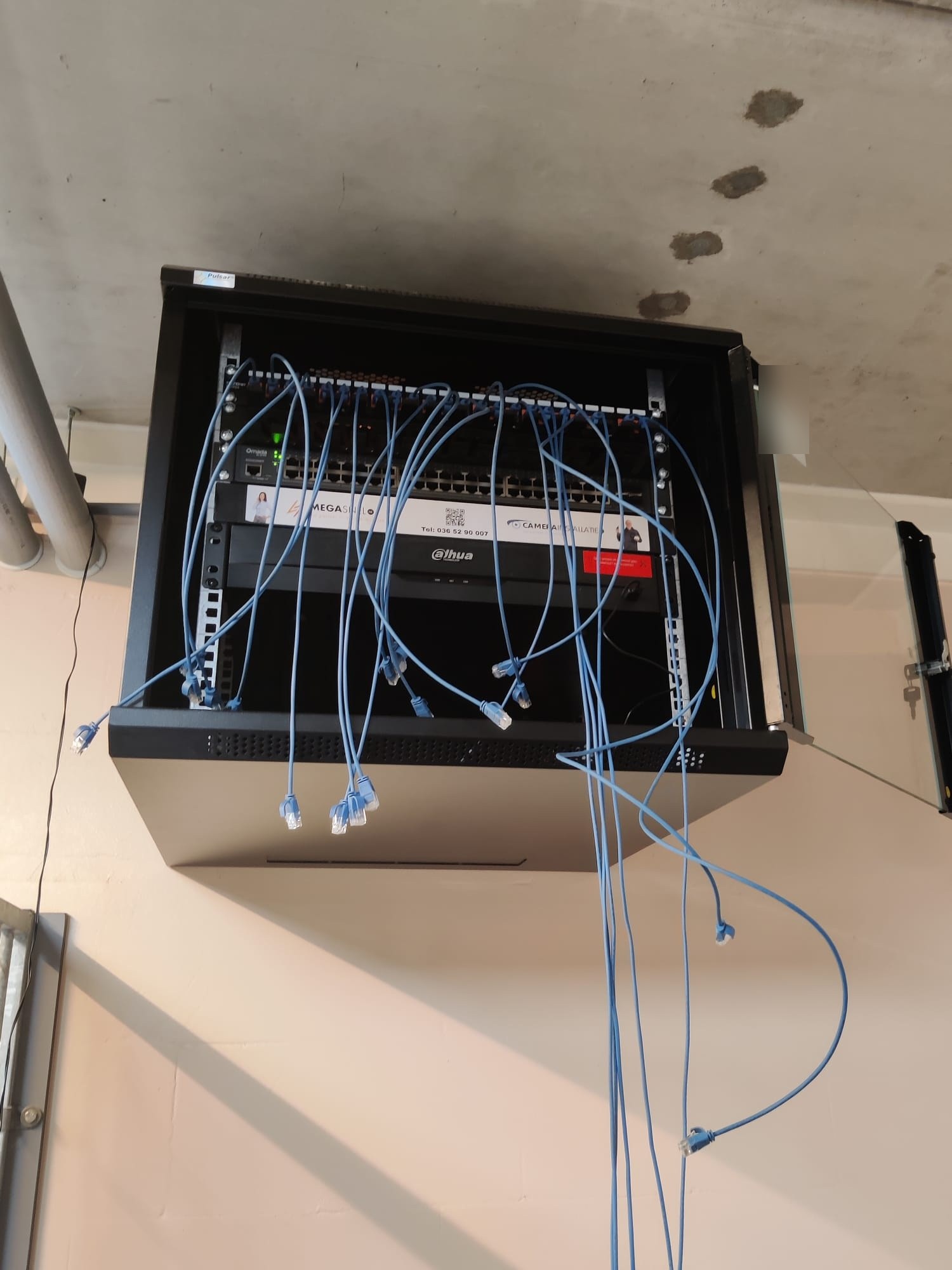 Open zwarte wandkast met patchpaneel, switch en recorder, met blauwe patchkabels die nog aangesloten worden