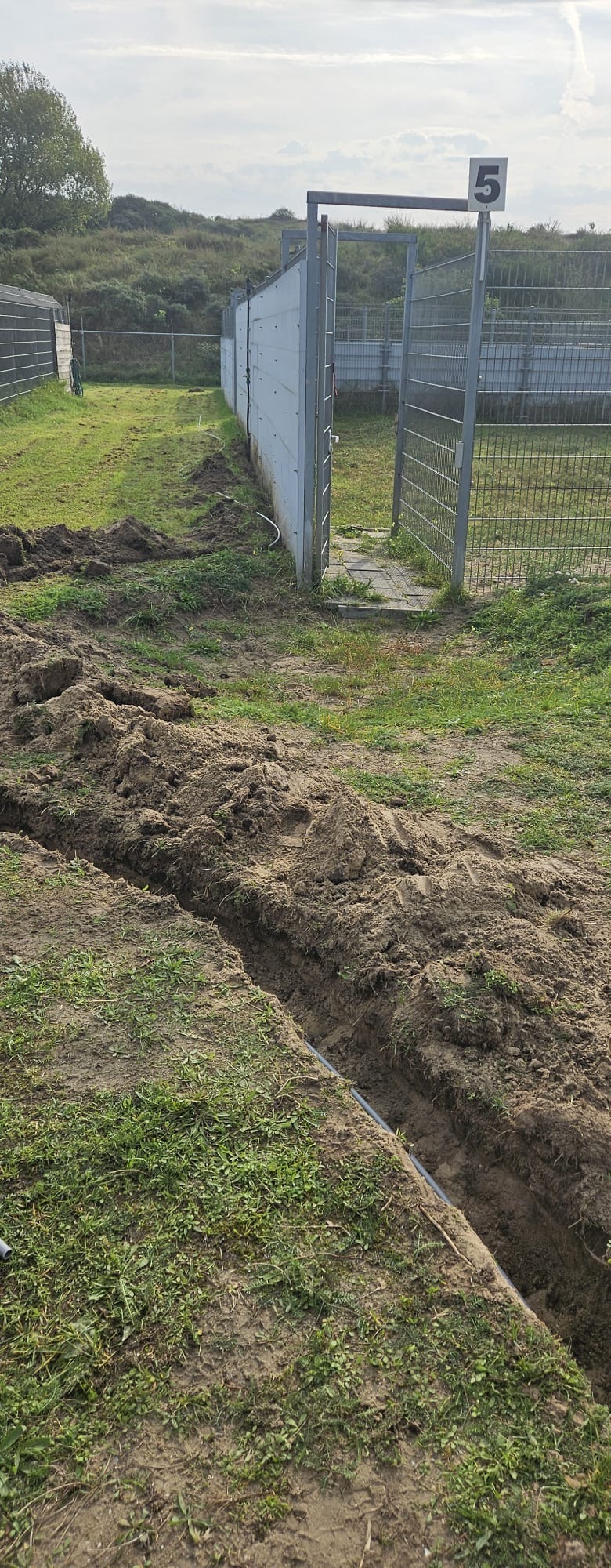 Open sleuf met grijze mantelbuis langs de buitenrennen, met een genummerd poortje en de duinen op de achtergrond