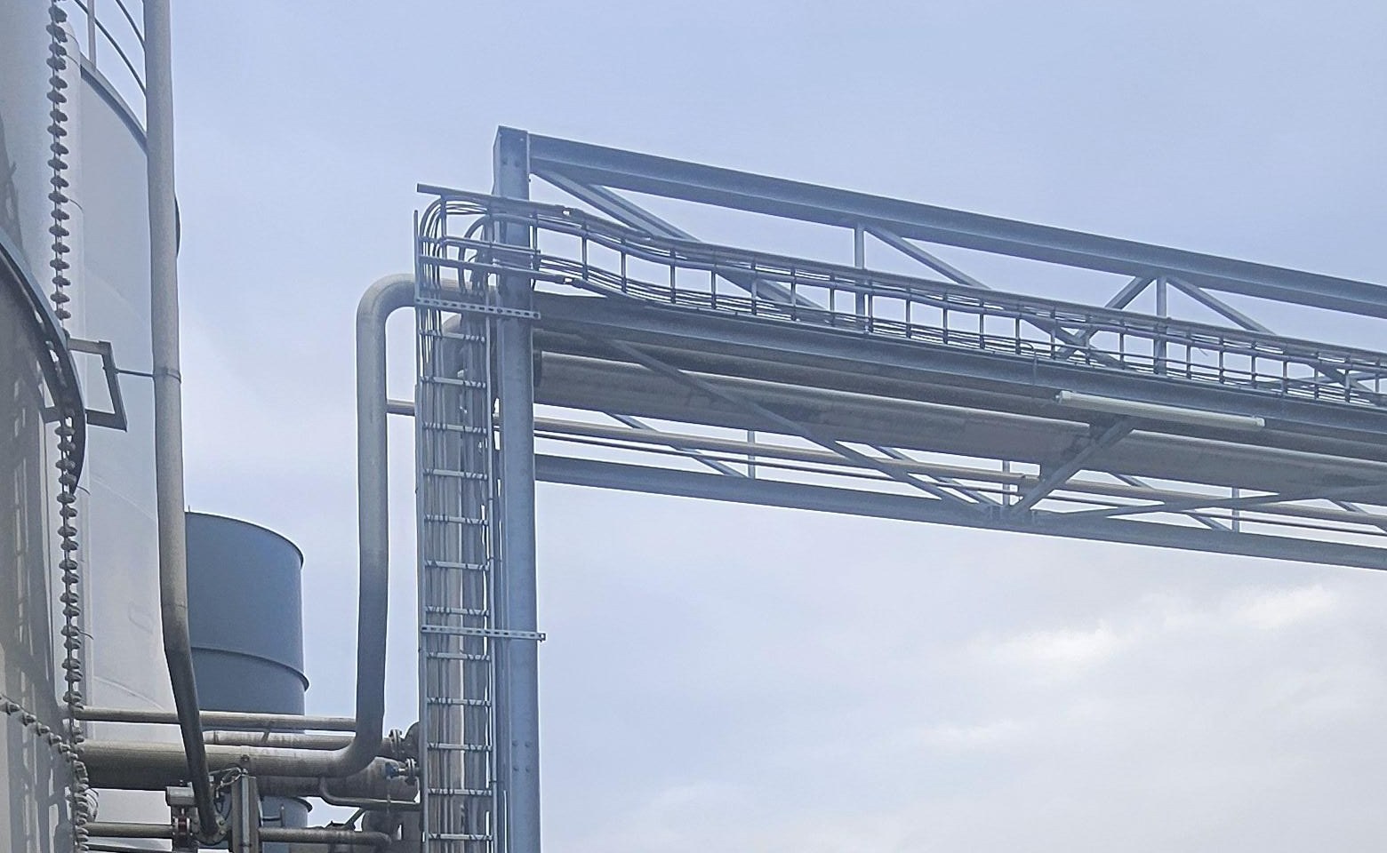 Stalen pijpbrug met loopbrug en ladder naast hoge opslagtanks, tegen een grijze lucht