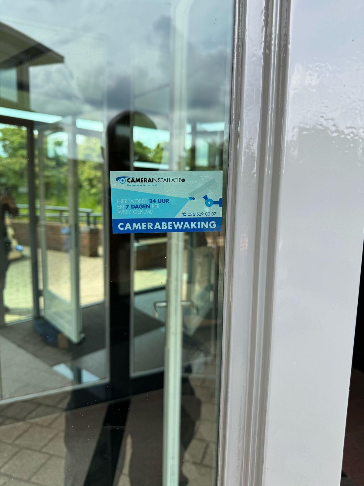 Sticker camerabewaking van CameraInstallatie.nl op een glazen deur, met de overkapping gespiegeld in het glas