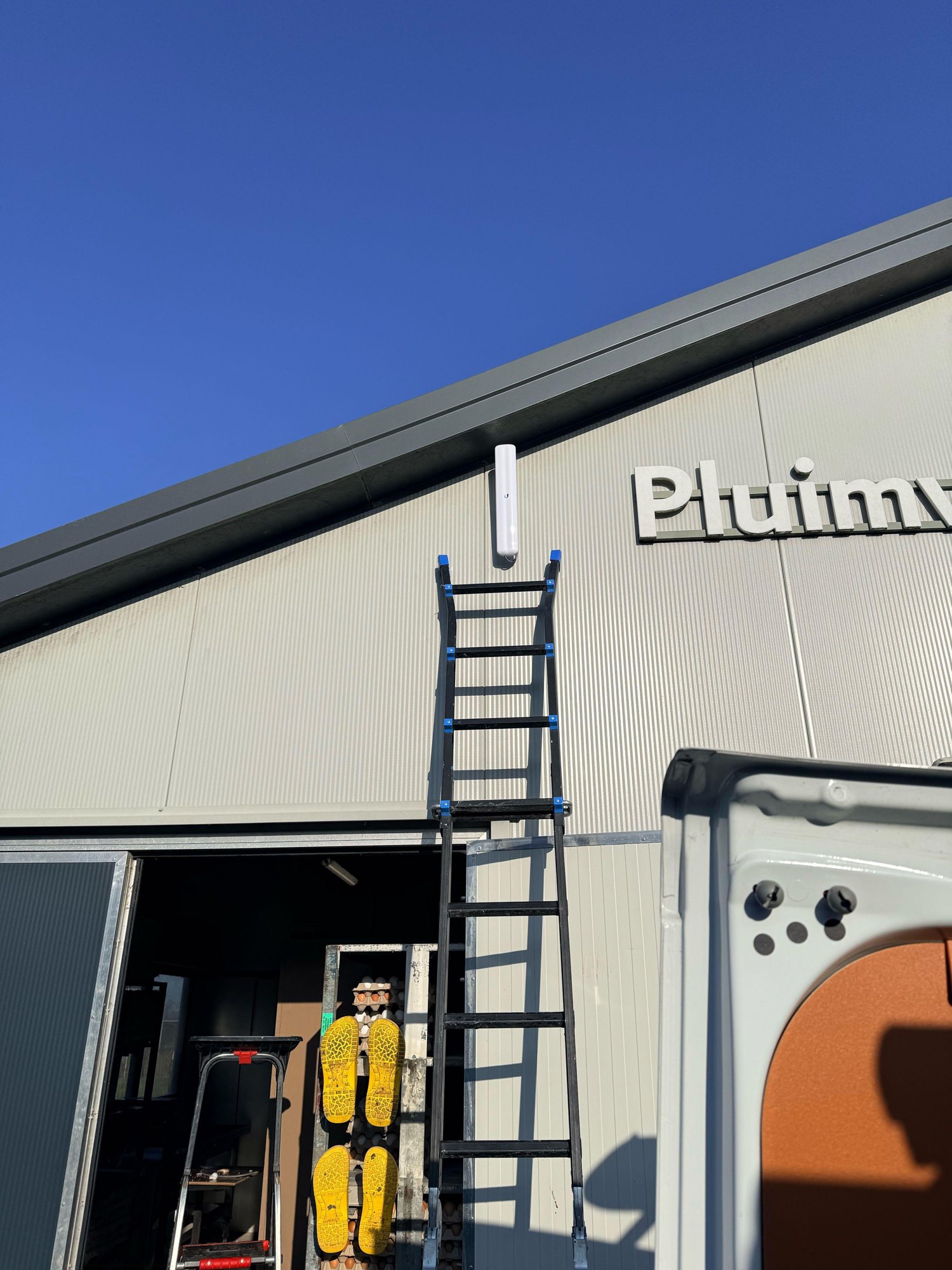 Witte straalzender hoog op de gevel van de stal onder de dakrand, met een ladder ertegen