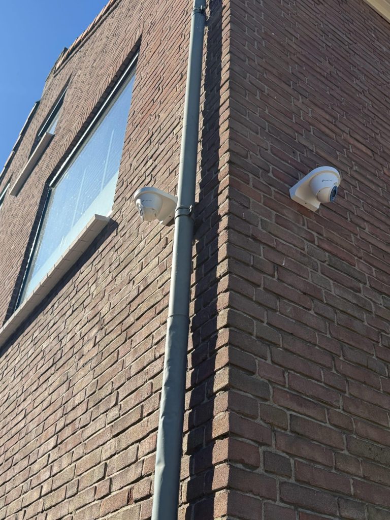 Twee witte VIGI-turretcamera's op de hoek van een bakstenen gevel, naast een regenpijp en onder een raam