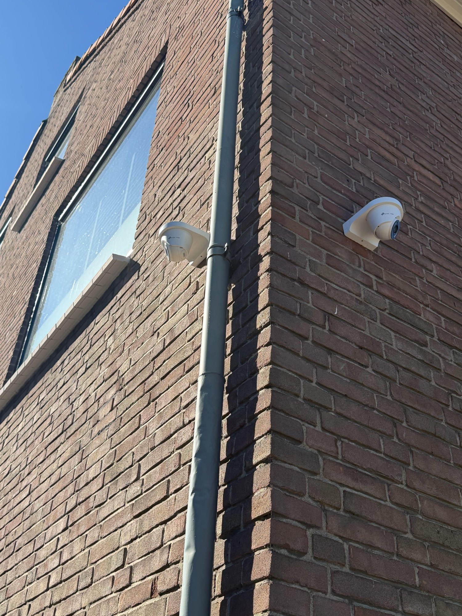 Twee witte VIGI-turretcamera's op de hoek van een bakstenen gevel, naast een regenpijp en onder een raam