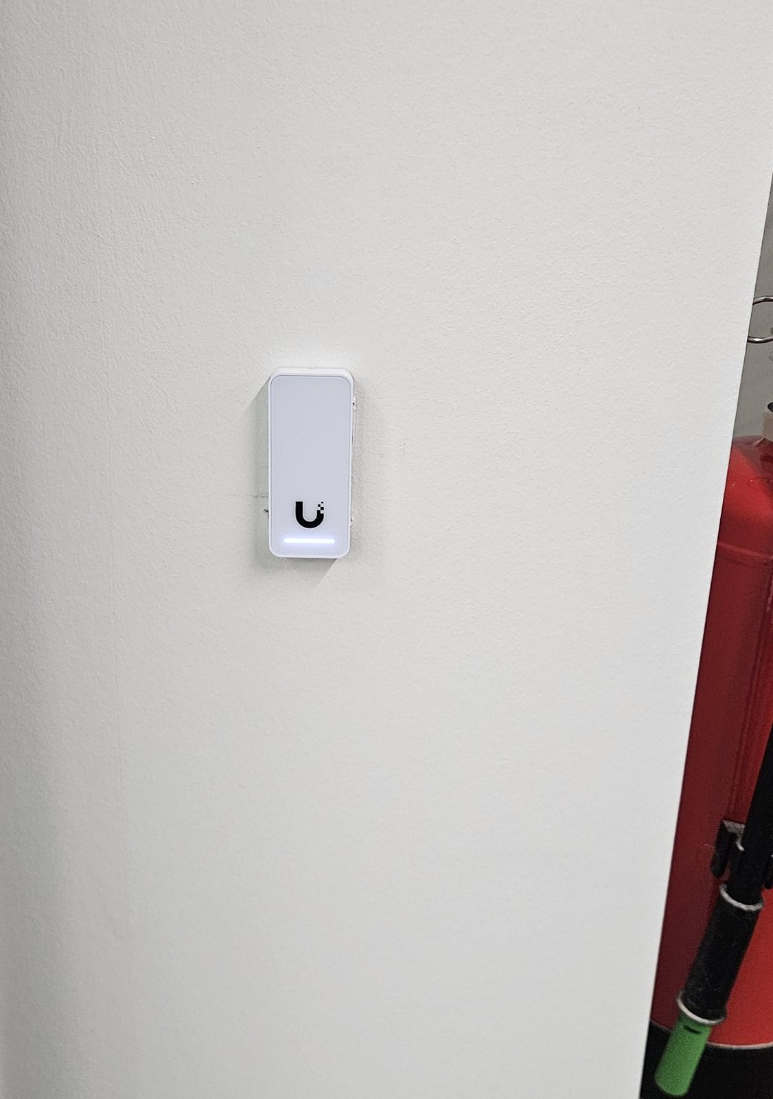 Witte UniFi-toegangslezer met blauw lampje op een witte muur naast een deur