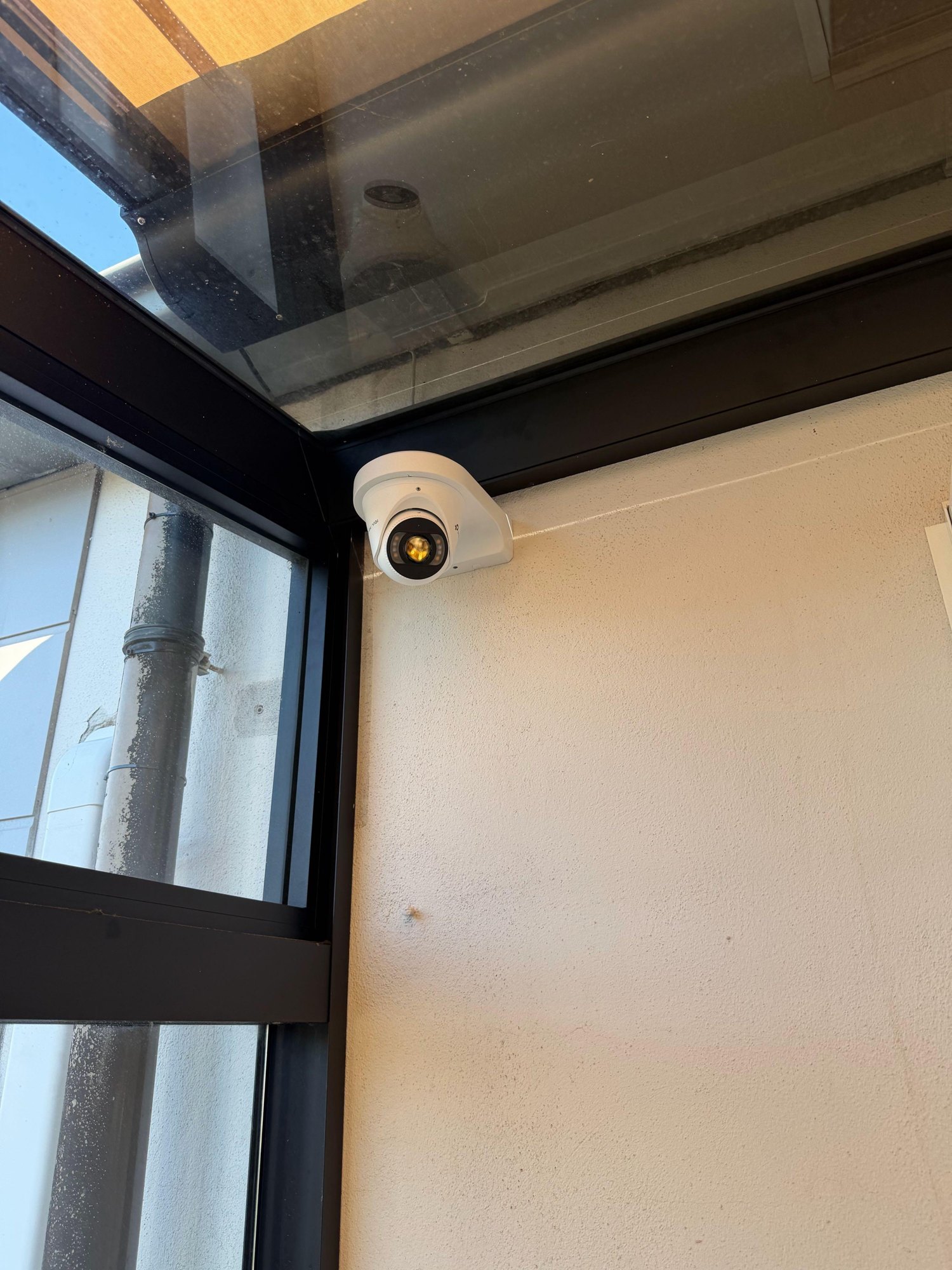 Witte VIGI-turretcamera met wandbeugel in de hoek onder een glazen overkapping, naast een zwart raamkozijn