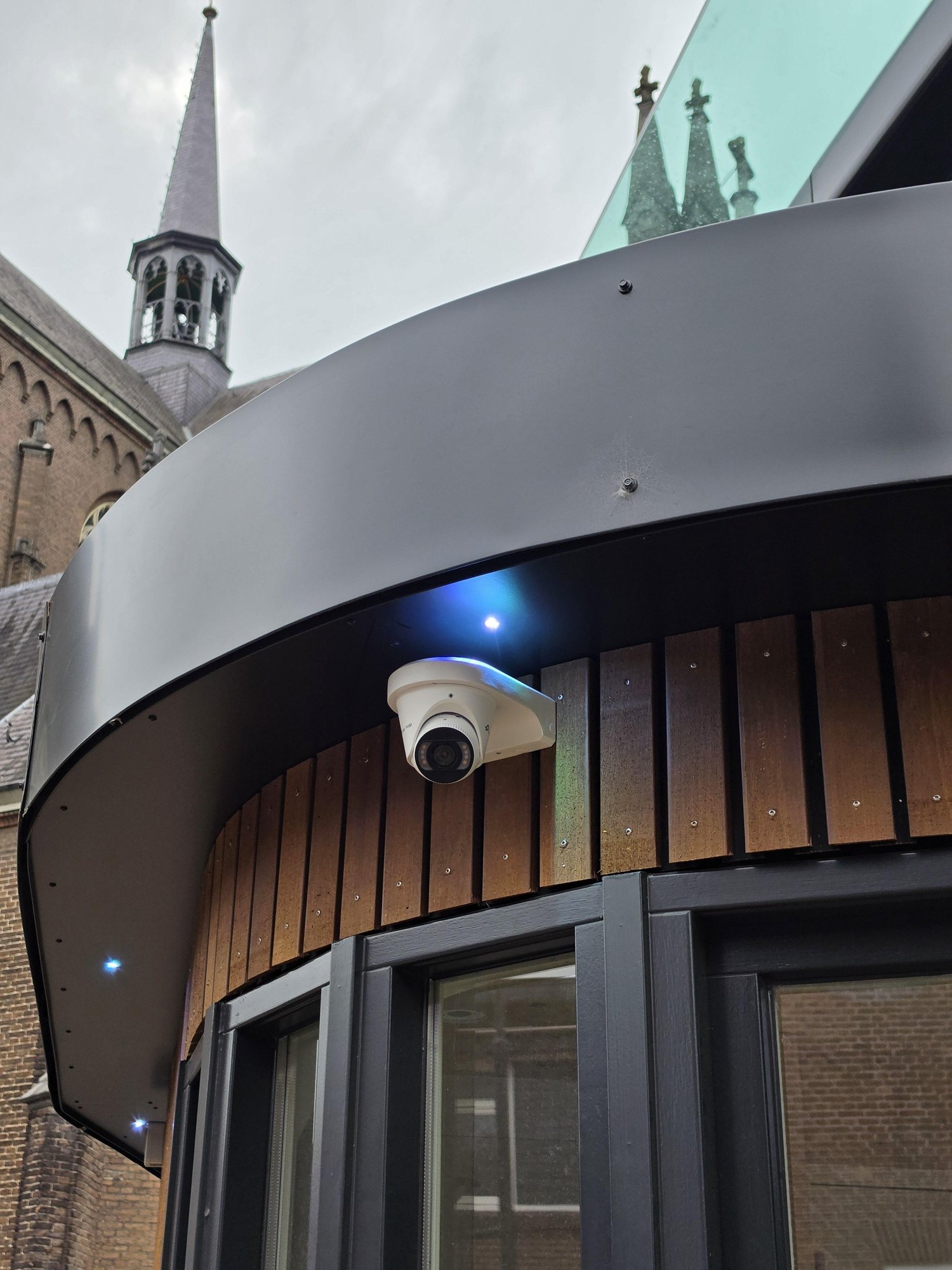 Witte VIGI-turretcamera onder de zwarte dakrand van het beheerdershuisje, met een kerktoren op de achtergrond