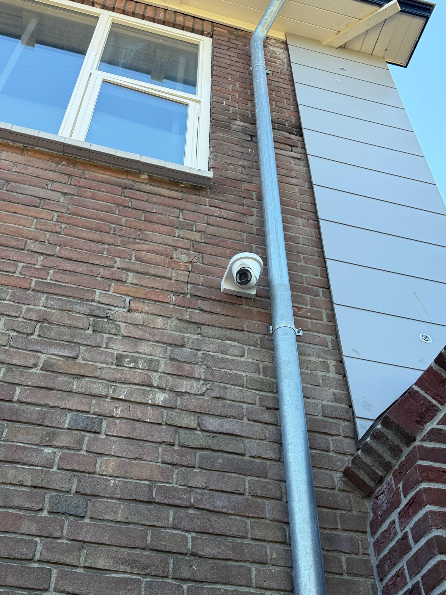 Witte VIGI-turretcamera op een bakstenen gevel naast een zinken regenpijp, met een raam erboven