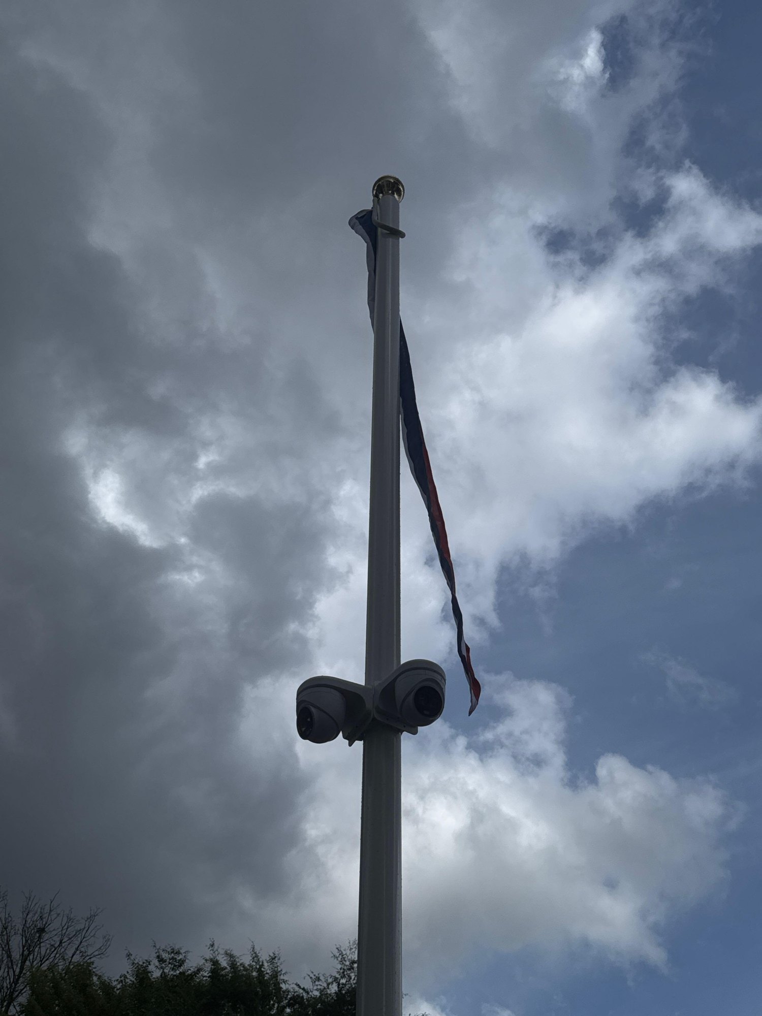 Witte vlaggenmast van onderen gezien, met halverwege twee camera’s en bovenin de vlag, tegen een wolkenlucht