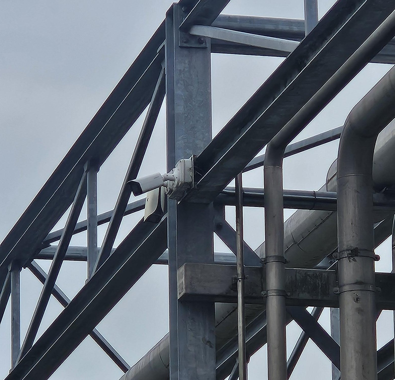 Witte bulletcamera aan een stalen staander van een pijpbrug, tussen leidingen