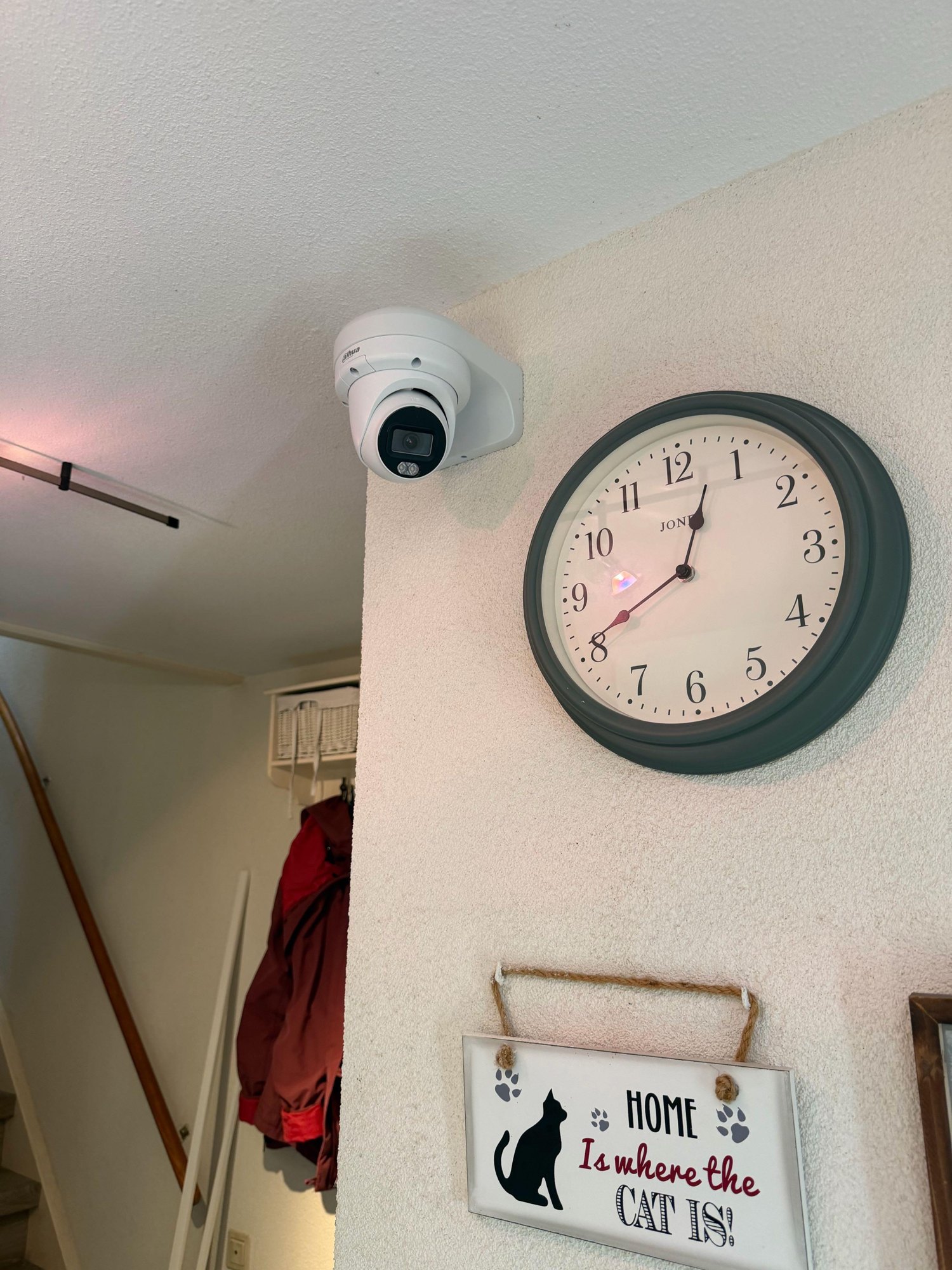 Witte camera op een witte beugel hoog aan de muur in een gang, naast een klok, met de trap op de achtergrond