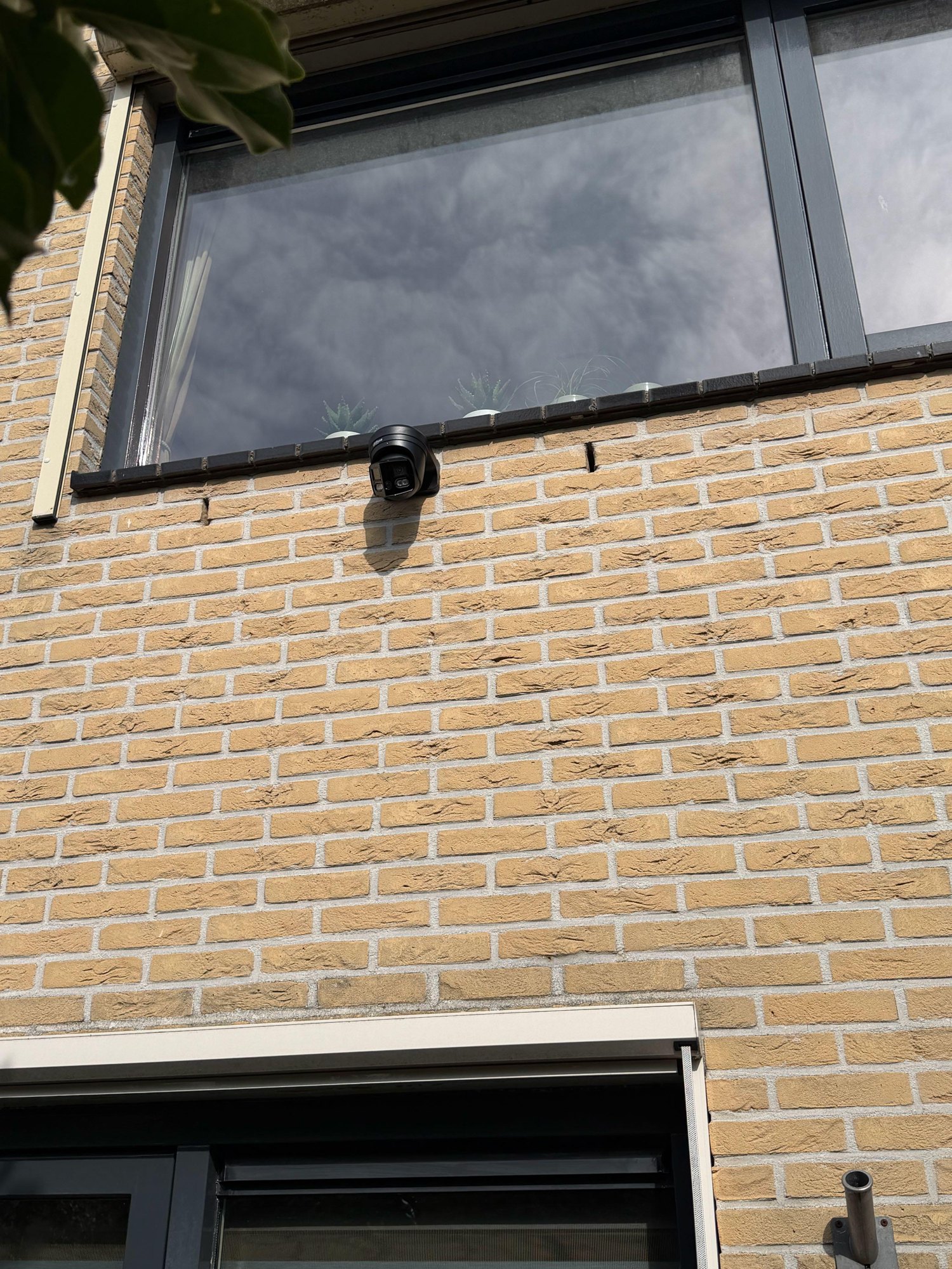 Zwarte 180 graden camera met twee lenzen op een gele bakstenen gevel, vlak onder een raam