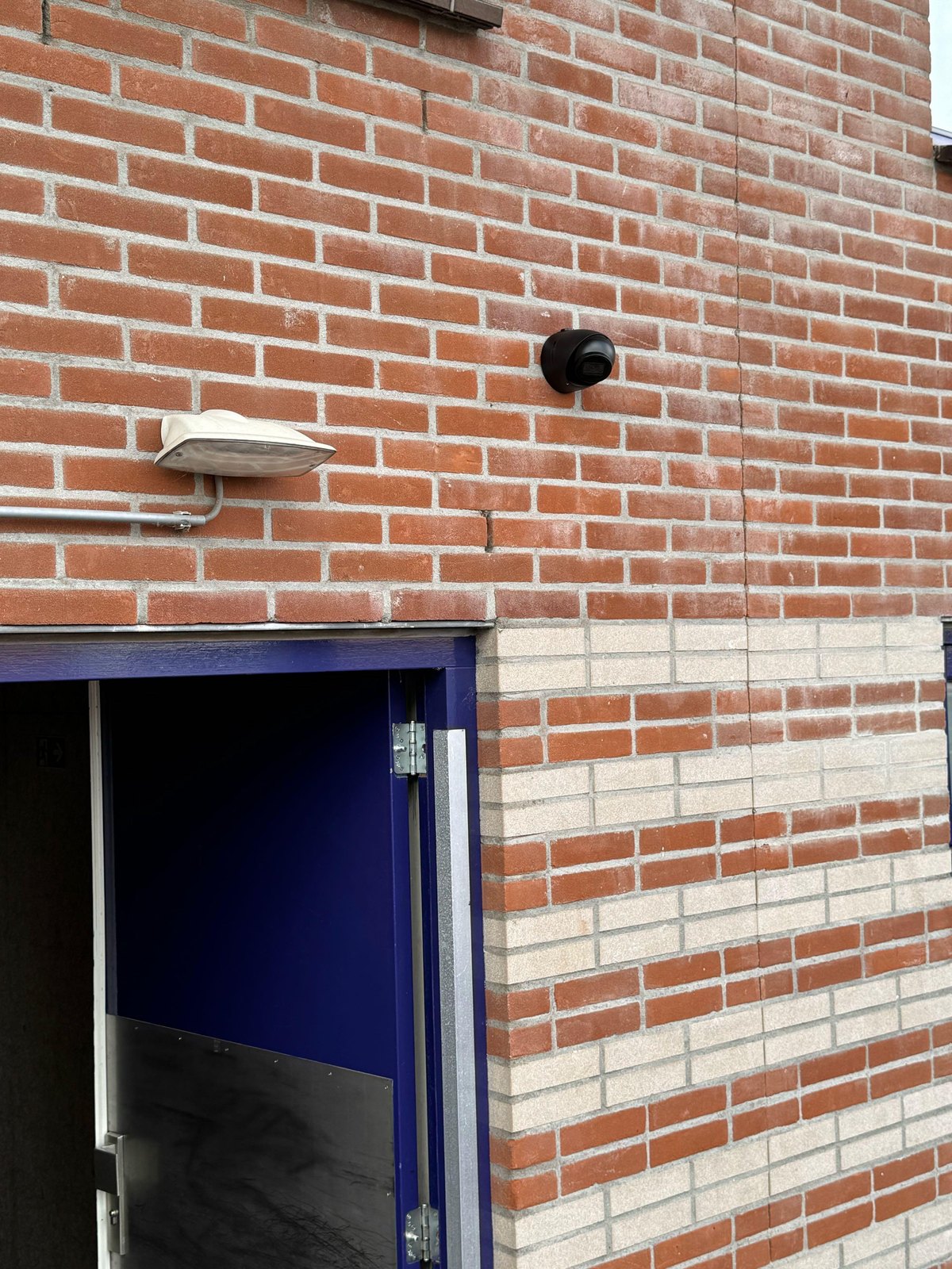 Zwarte camera op een bakstenen muur boven een blauwe nooddeur, naast een buitenlamp