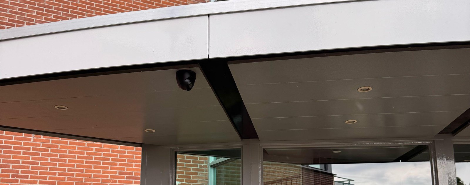 Zwarte camera op de hoek onder een aluminium overkapping, boven een glazen entree met bakstenen gevel