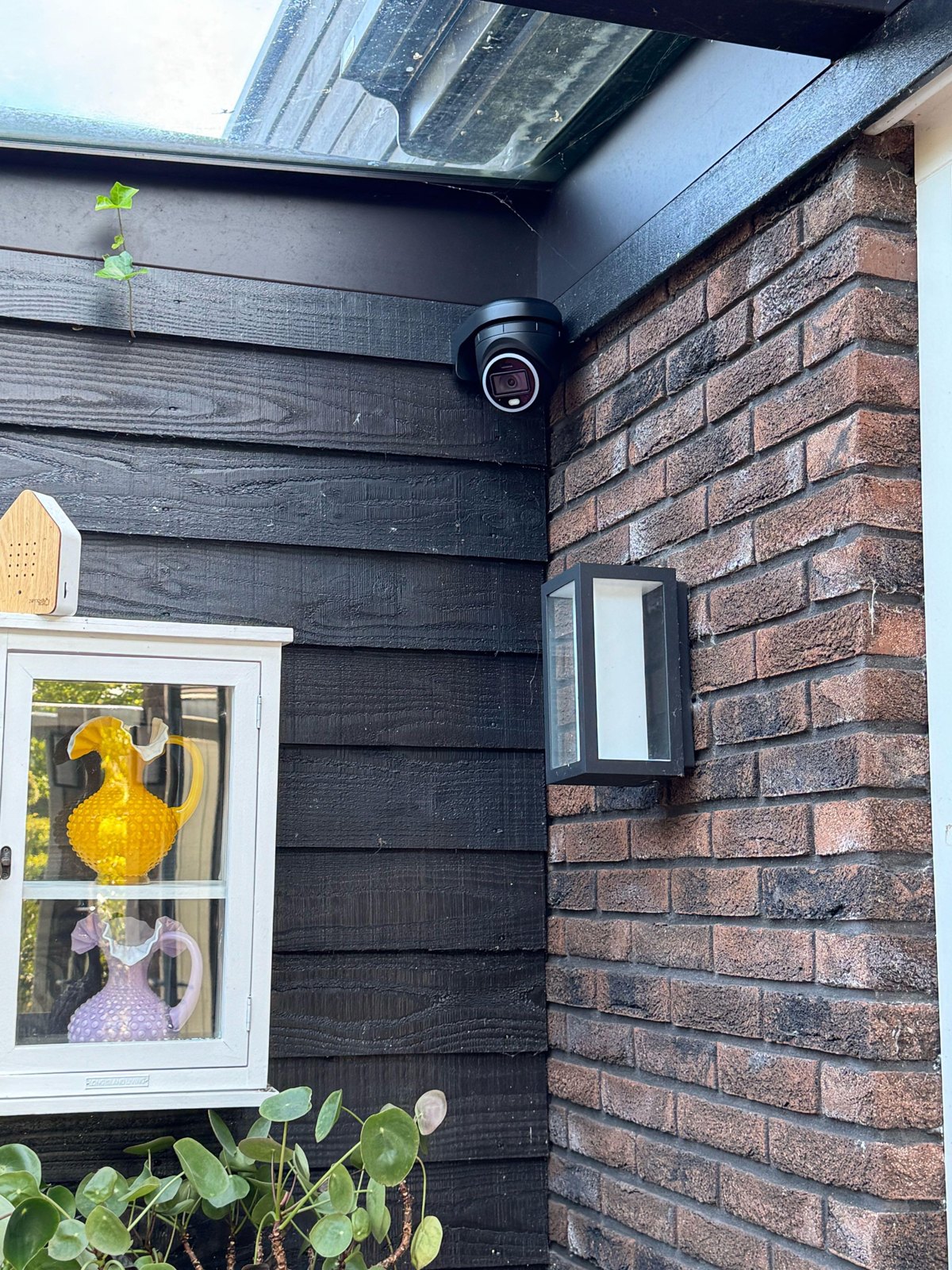 Zwarte turretcamera in de hoek tussen een zwart houten gevel en een bakstenen muur, naast een buitenlamp