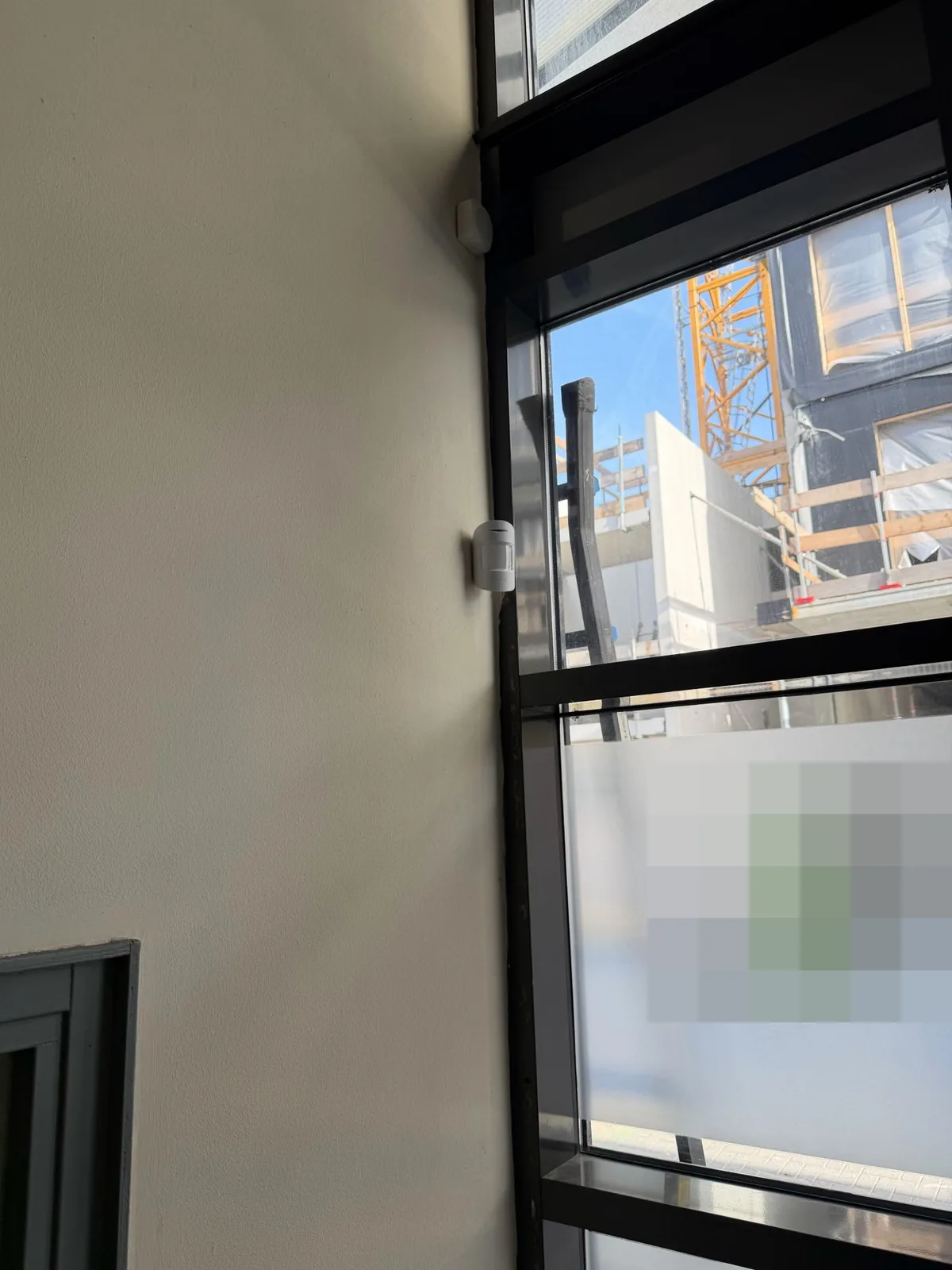Witte Ajax-bewegingsmelder op de wand naast een glazen pui, met buiten een bouwkraan