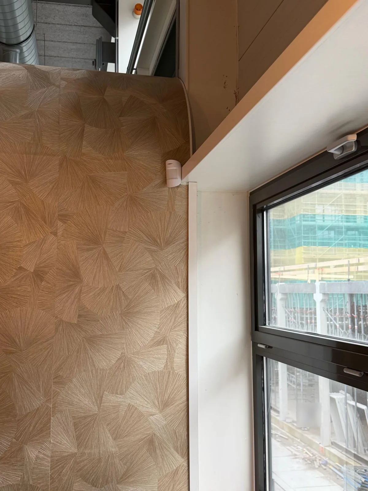 Witte Ajax-bewegingsmelder op de wand naast een raam, tegen een houten wandbekleding, met buiten een bouwplaats
