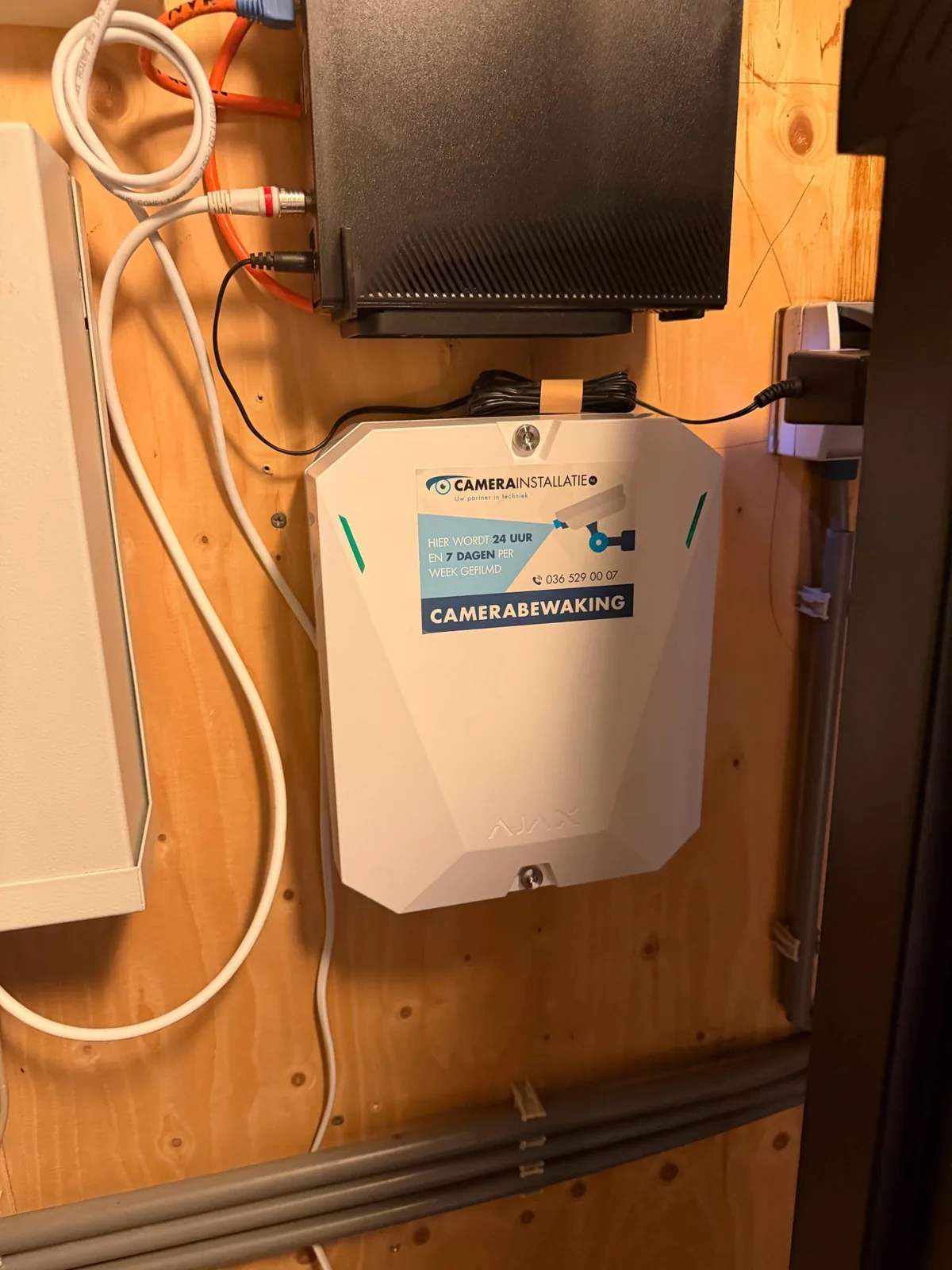 Witte Ajax Hub Hybrid voor bedrade Fibra-melders, met de sticker van CameraInstallatie.nl, op een houten wand