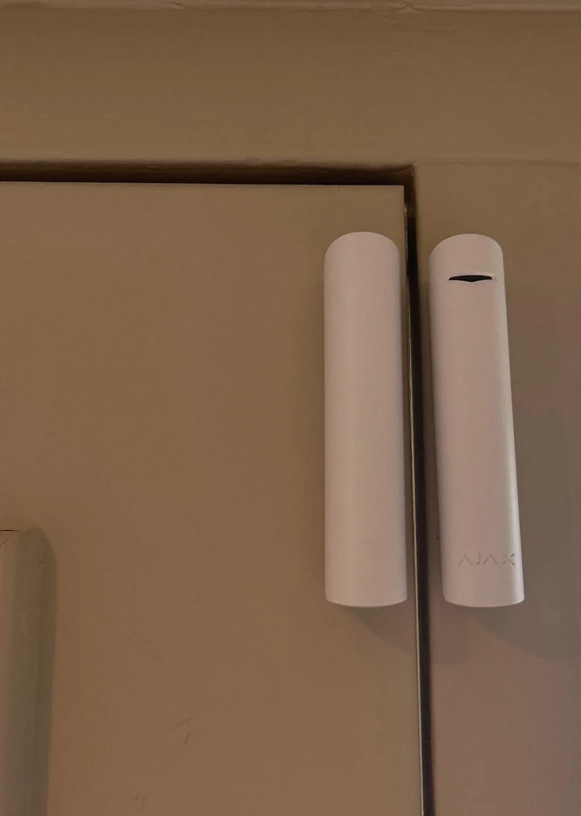 Wit Ajax-magneetcontact Superior DoorProtect bovenin een kozijn: rechts de melder met het Ajax-logo, links de magneet op het deel dat opengaat