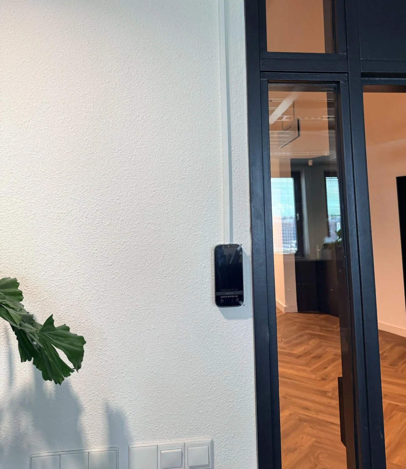 Zwart Ajax-bediendeel met touchscreen op een witte wand naast een zwarte glazen deur, bij de entree van een kantoor in Amsterdam