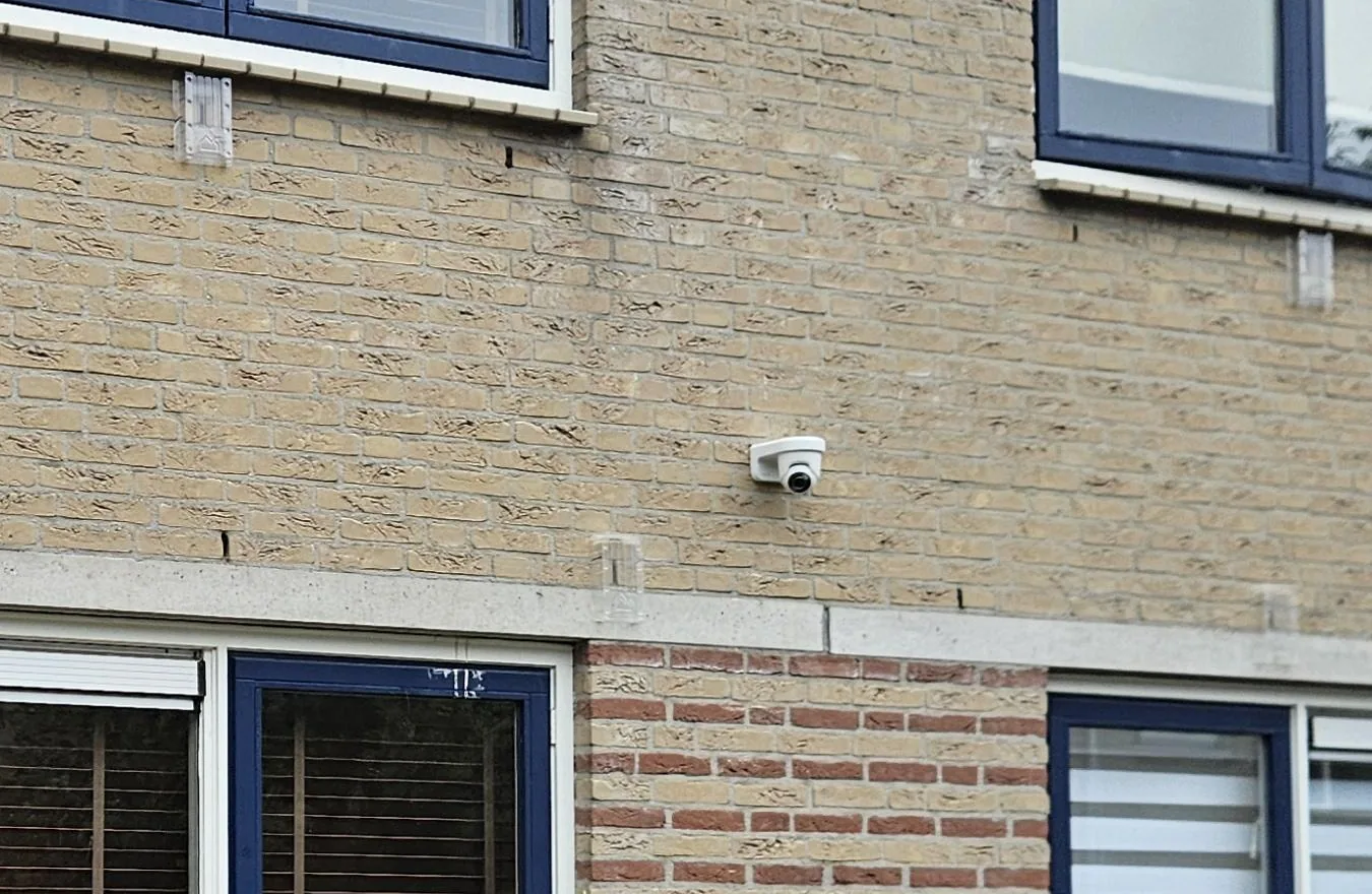 Witte Ajax TurretCam op een muurbeugel tegen de gele bakstenen voorgevel, tussen de ramen van de begane grond en de eerste verdieping