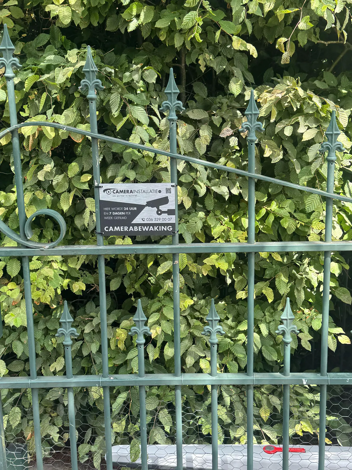 Bord camerabewaking van CameraInstallatie.nl aan een groen smeedijzeren hek met een haag erachter