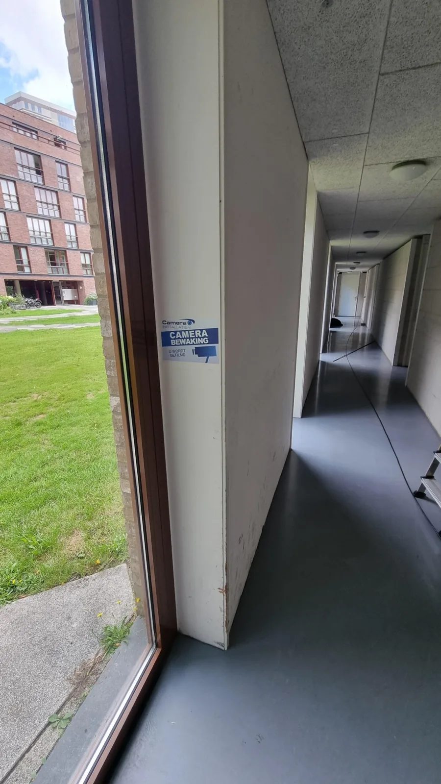 Sticker camerabewaking op het glas bij de ingang, met de gang erachter