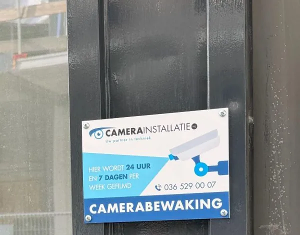 Bord van CameraInstallatie.nl met de tekst camerabewaking op het donkere kozijn van de entree