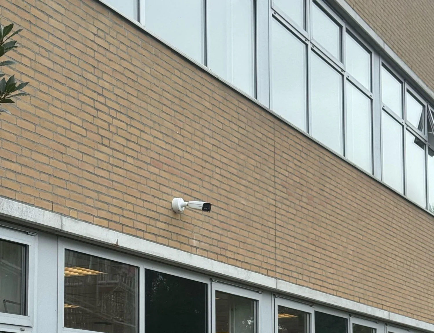 Witte bulletcamera hoog op een gevel van gele baksteen, boven een rij ramen