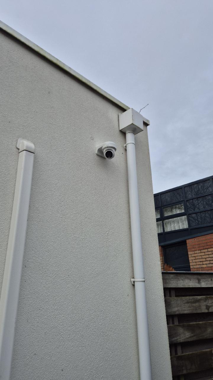 Witte camera op een witte gevel tussen twee witte regenpijpen, onder een grijze lucht