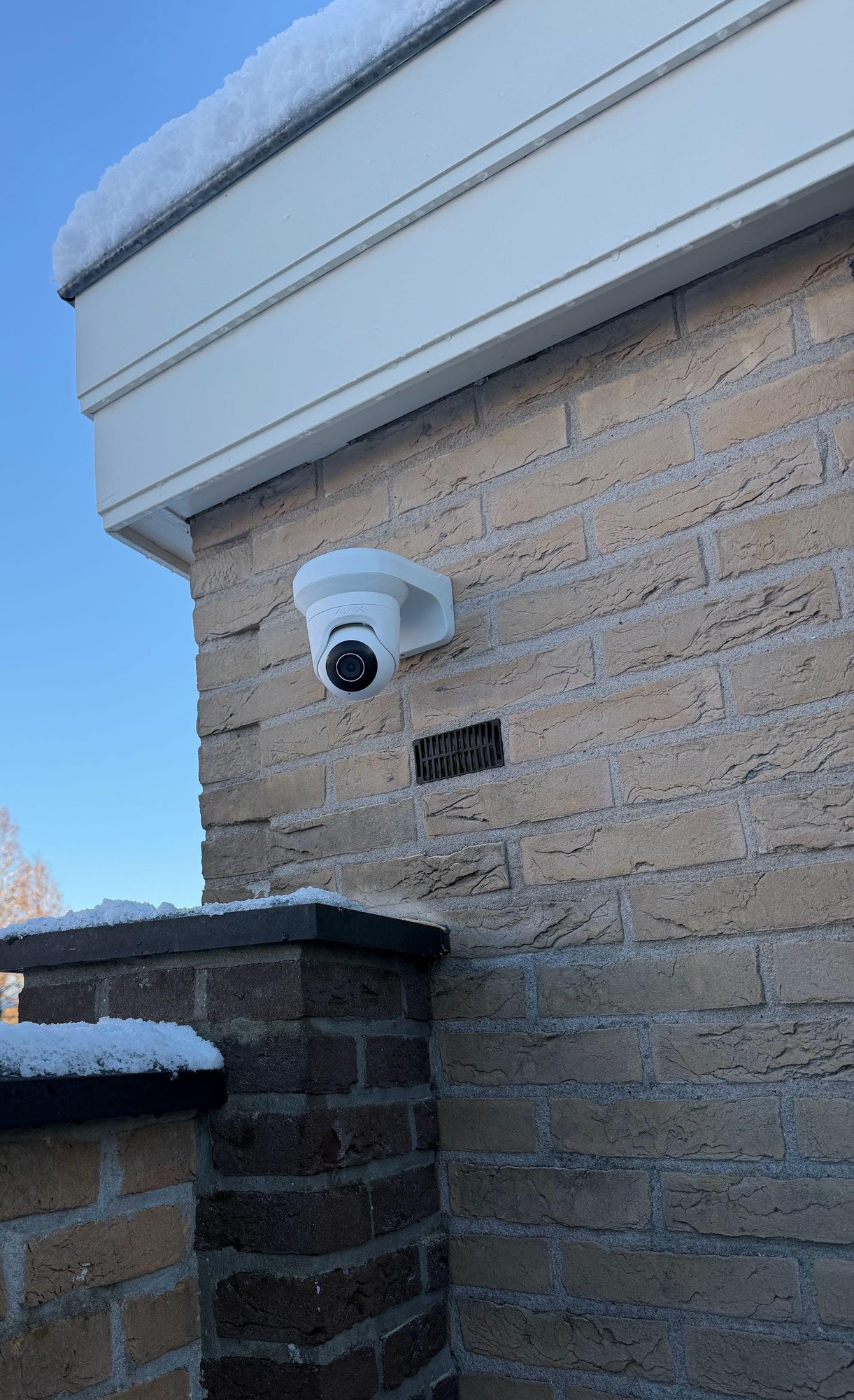 Witte camera hoog op de bakstenen zijgevel onder de dakrand, met sneeuw op de tuinmuur