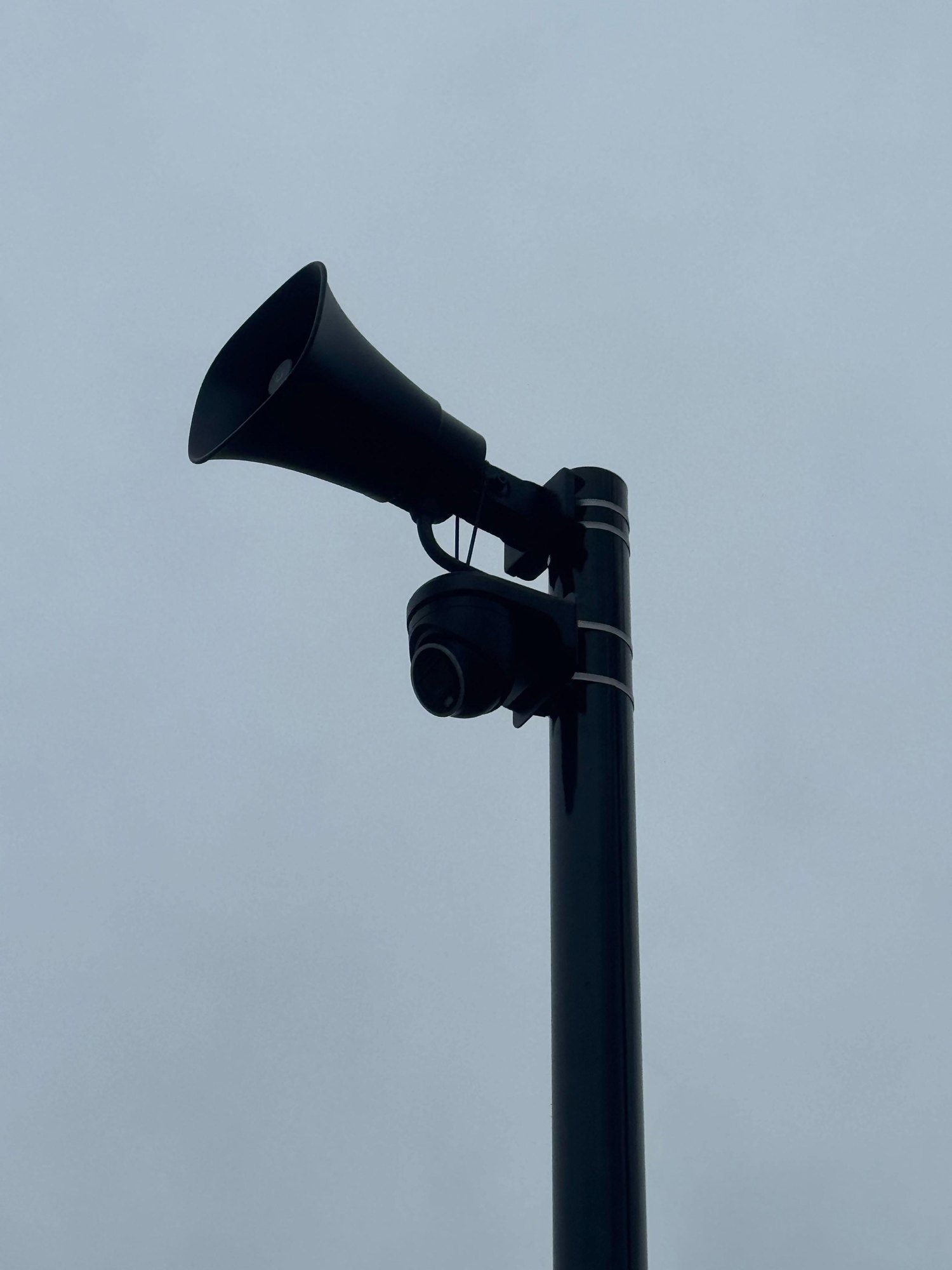 Zwarte hoorn en zwarte camera bovenaan een cameramast, tegen een grijze lucht