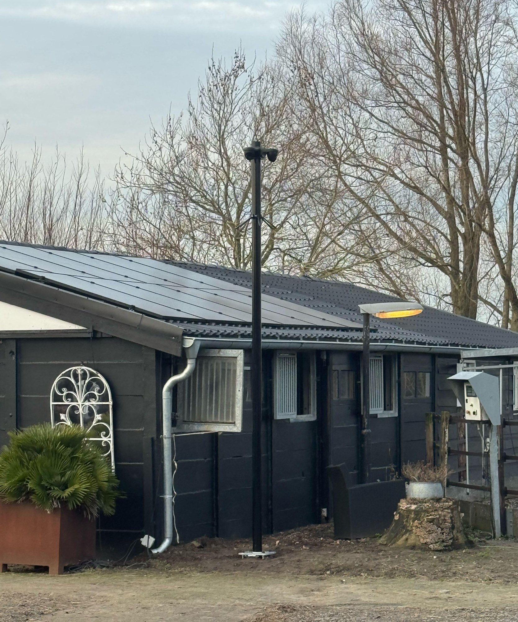Zwarte cameramast van 4 meter naast een zwarte paardenstal met zonnepanelen, kale bomen erachter