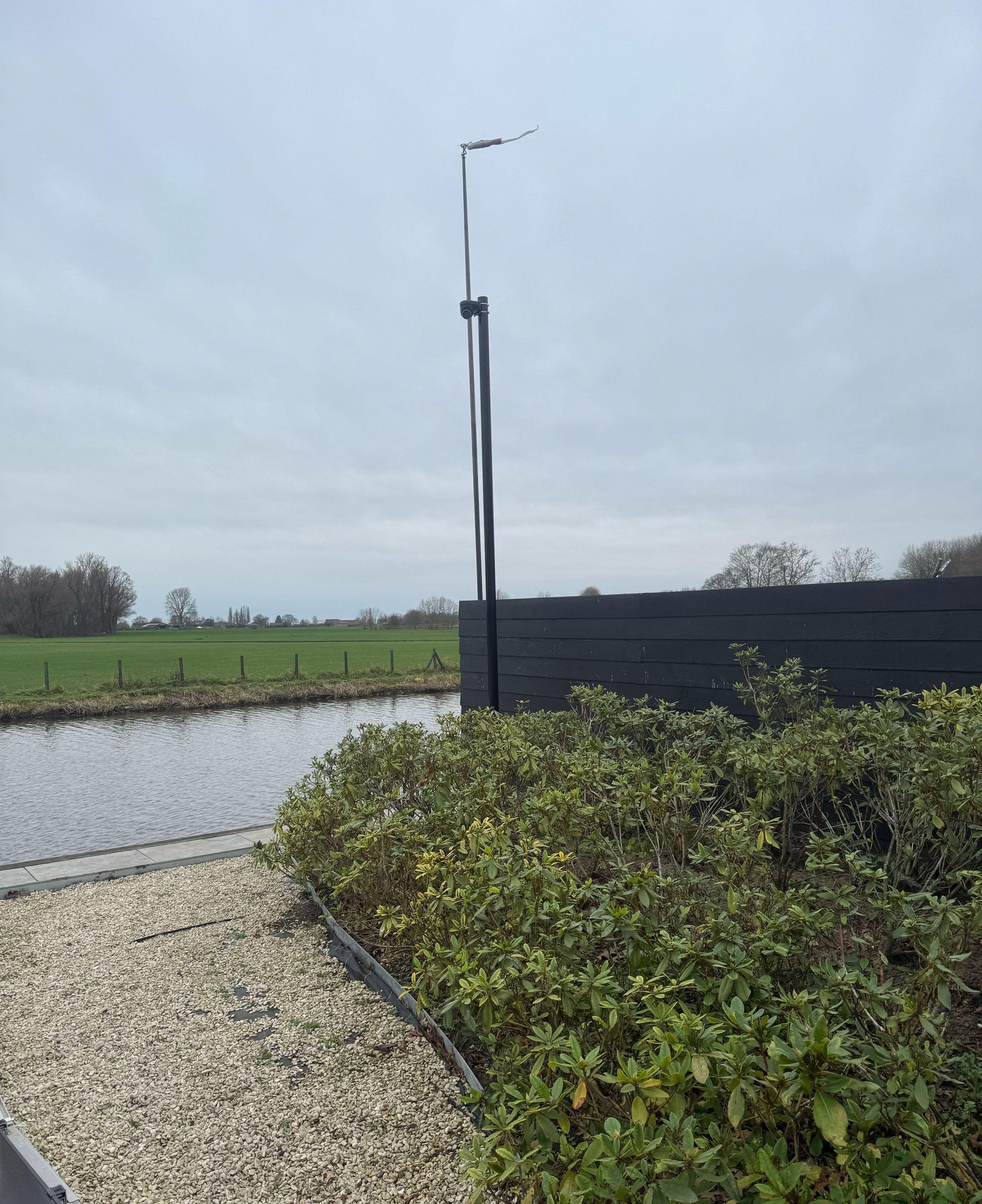 Zwarte cameramast van 4,5 meter achter in de tuin, naast een sloot met weiland erachter