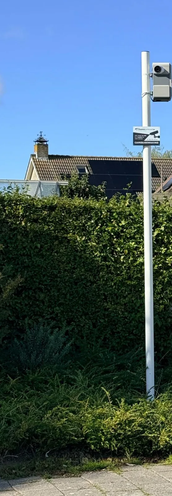 Witte camerapaal met kast en camera naast een hoge haag bij de poort van een bedrijfsterrein