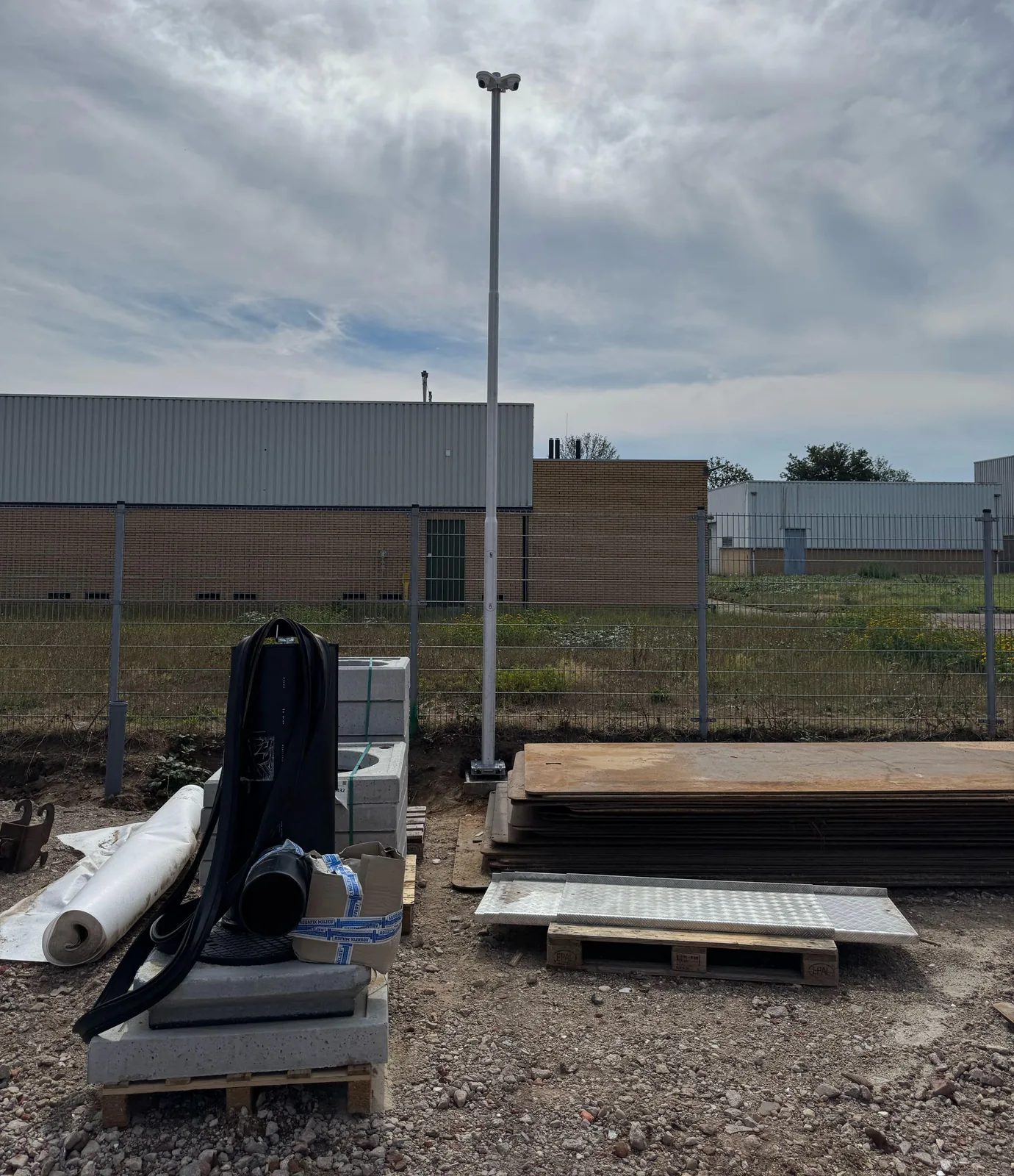 Hoge witte camerapaal met twee witte camera's op een T-beugel, vlak achter een gaashekwerk, met bouwmaterialen en rijplaten op de voorgrond en loodsen erachter