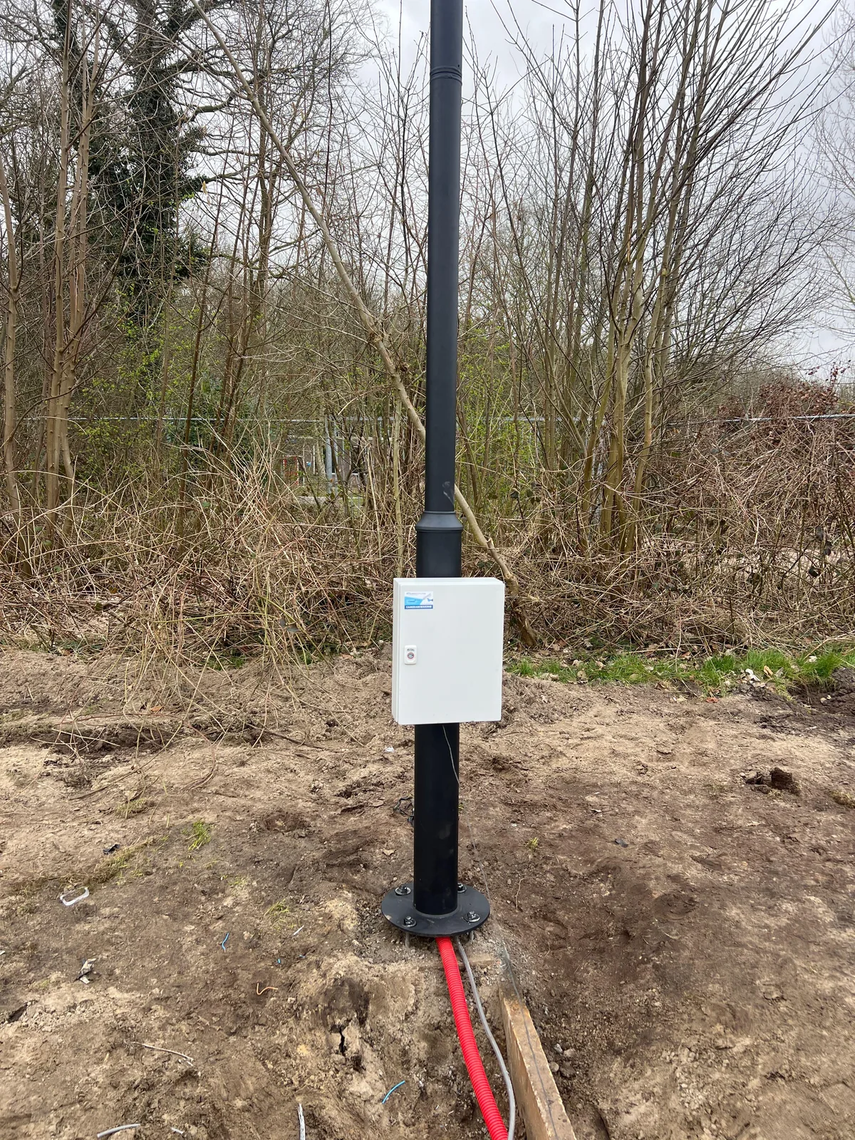 Zwarte paal op een bestaande fundering met een witte kast, de rode mantelbuis met glasvezel gaat onder de paal in