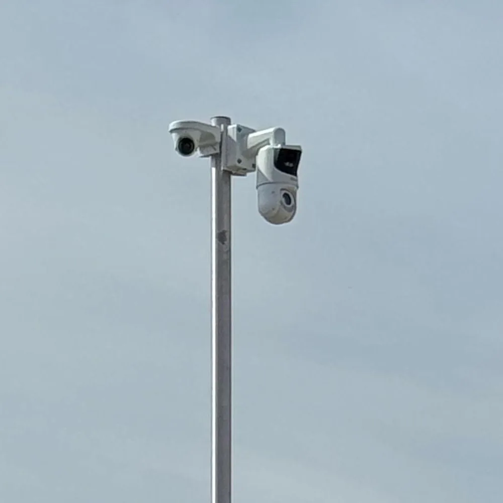 Bovenkant van een camerapaal tegen een grijze lucht: links een kleine witte camera op een beugel, rechts een witte Dahua dual-lens PTZ met een brede lens boven de draaibare koepel