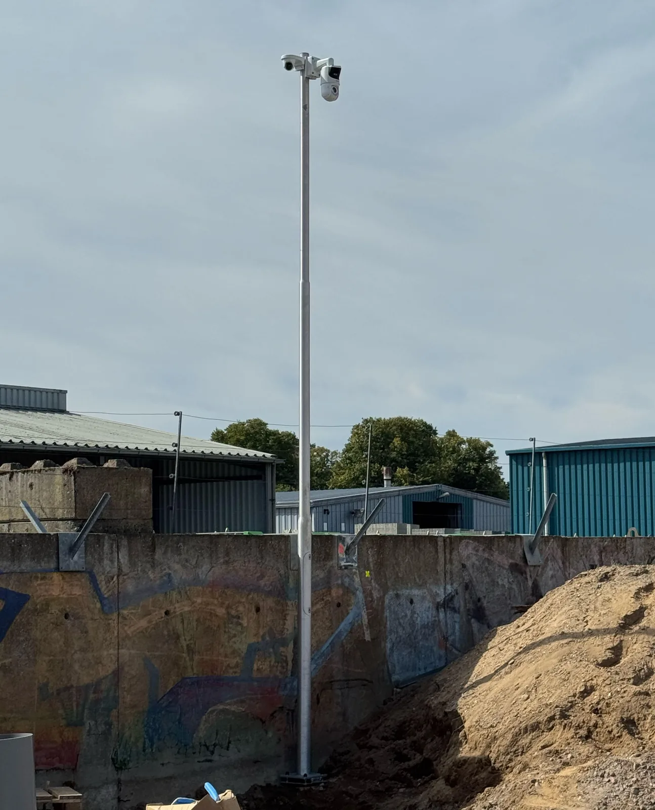 Verzinkte camerapaal voor een betonnen keerwand met graffiti, bovenop een witte Dahua dual-lens PTZ-camera en een kleinere camera op een beugel, ernaast een hoop zand en loodsen op de achtergrond