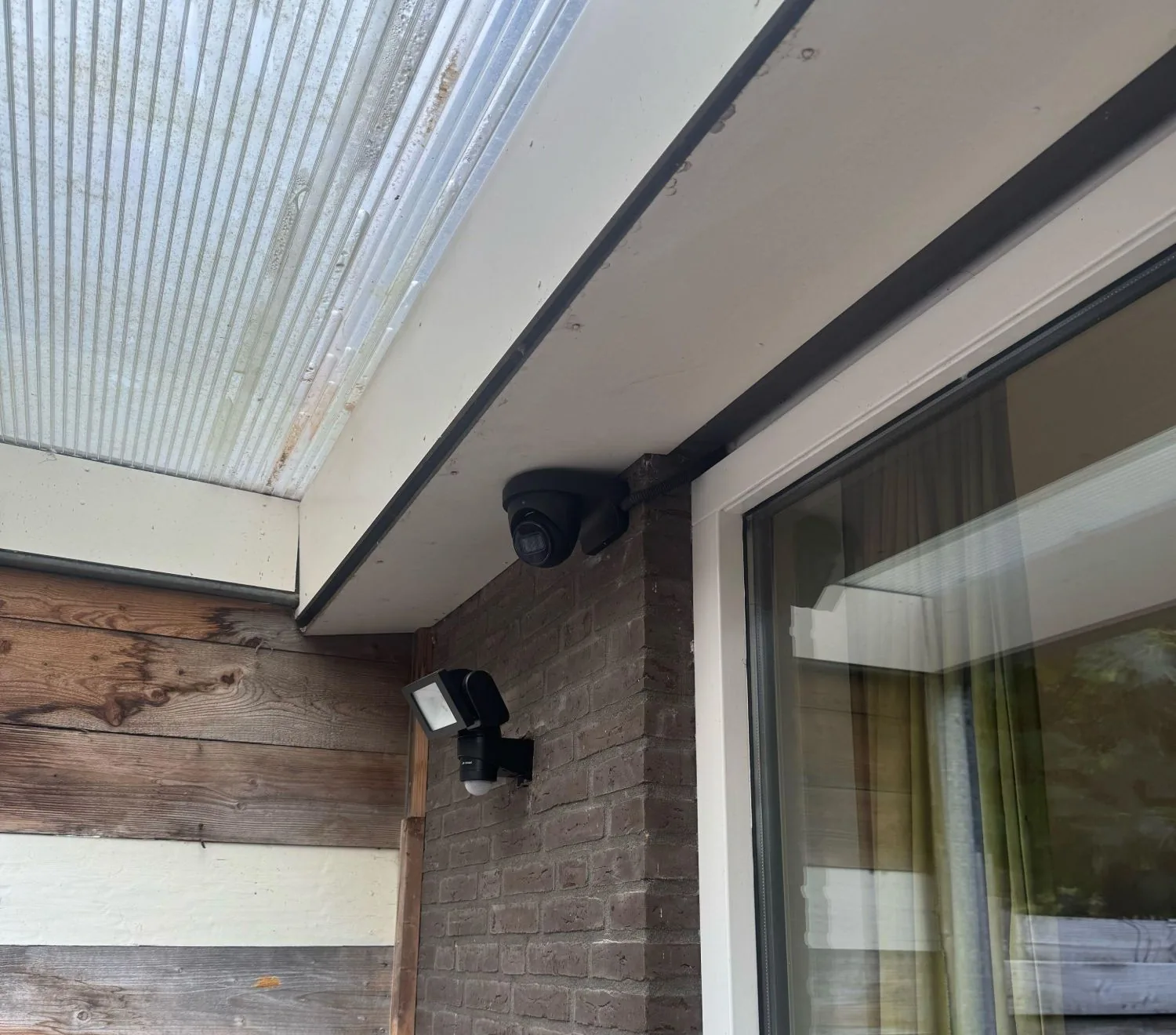Zwarte Dahua-turretcamera onder een overkapping, op de hoek van de gevel naast een schuifpui, met een buitenlamp met bewegingsmelder eronder