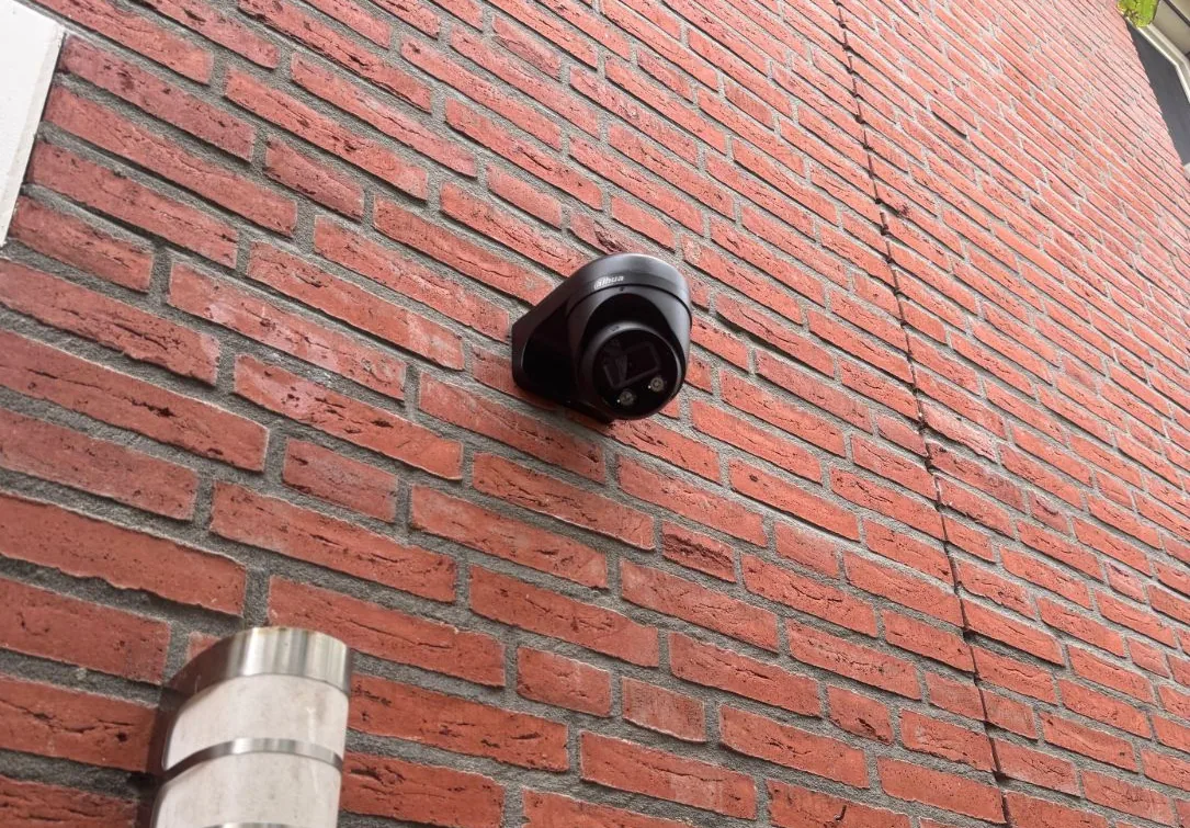 Zwarte Dahua TiOC-camera op een muurbeugel tegen een rode bakstenen voorgevel, met een buitenlamp eronder