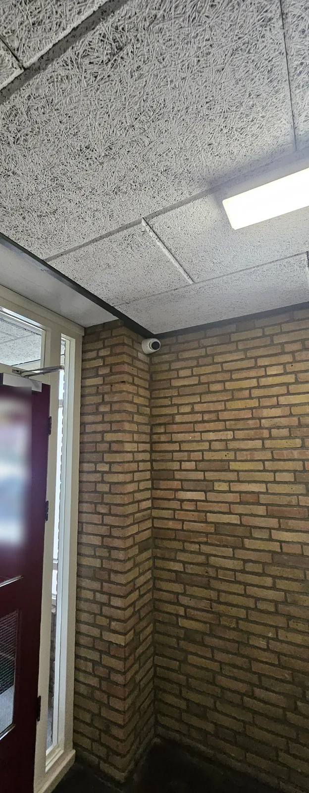 Entreehal met gele bakstenen muren en een houtwolplafond, met hoog in de hoek een witte camera naast een donkerrode deur