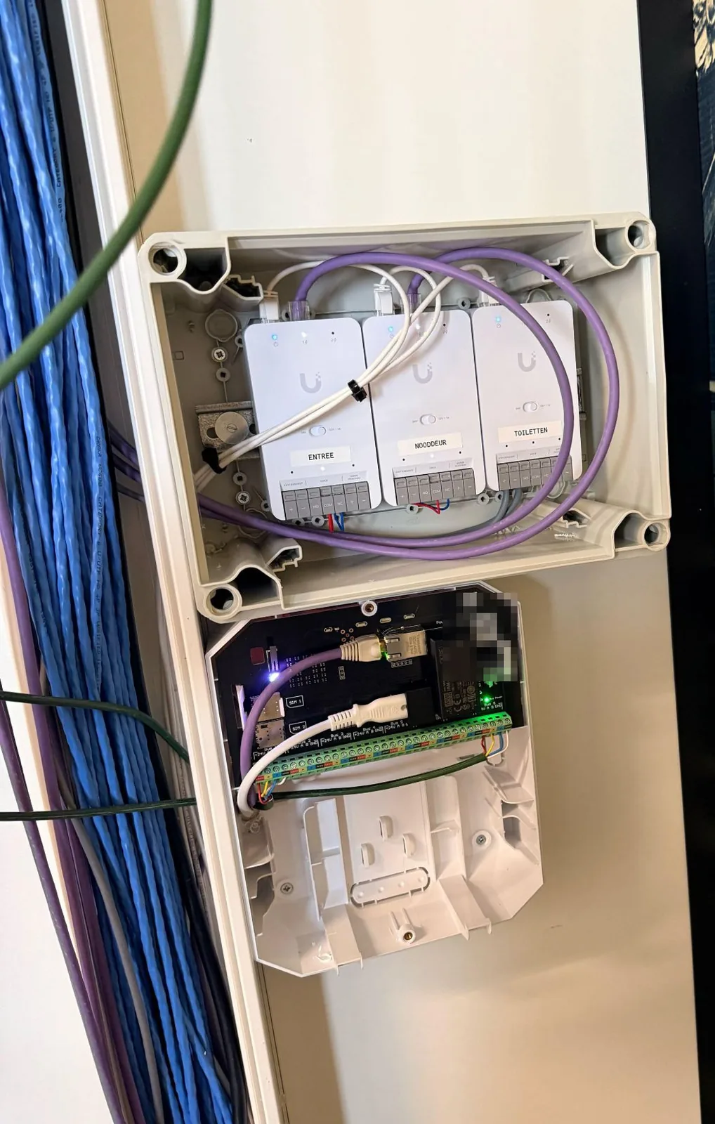 Bak met drie UniFi Door Hub Mini's met de labels Entree, Nooddeur en Toiletten, daaronder de open Ajax-hub met aangesloten bussen