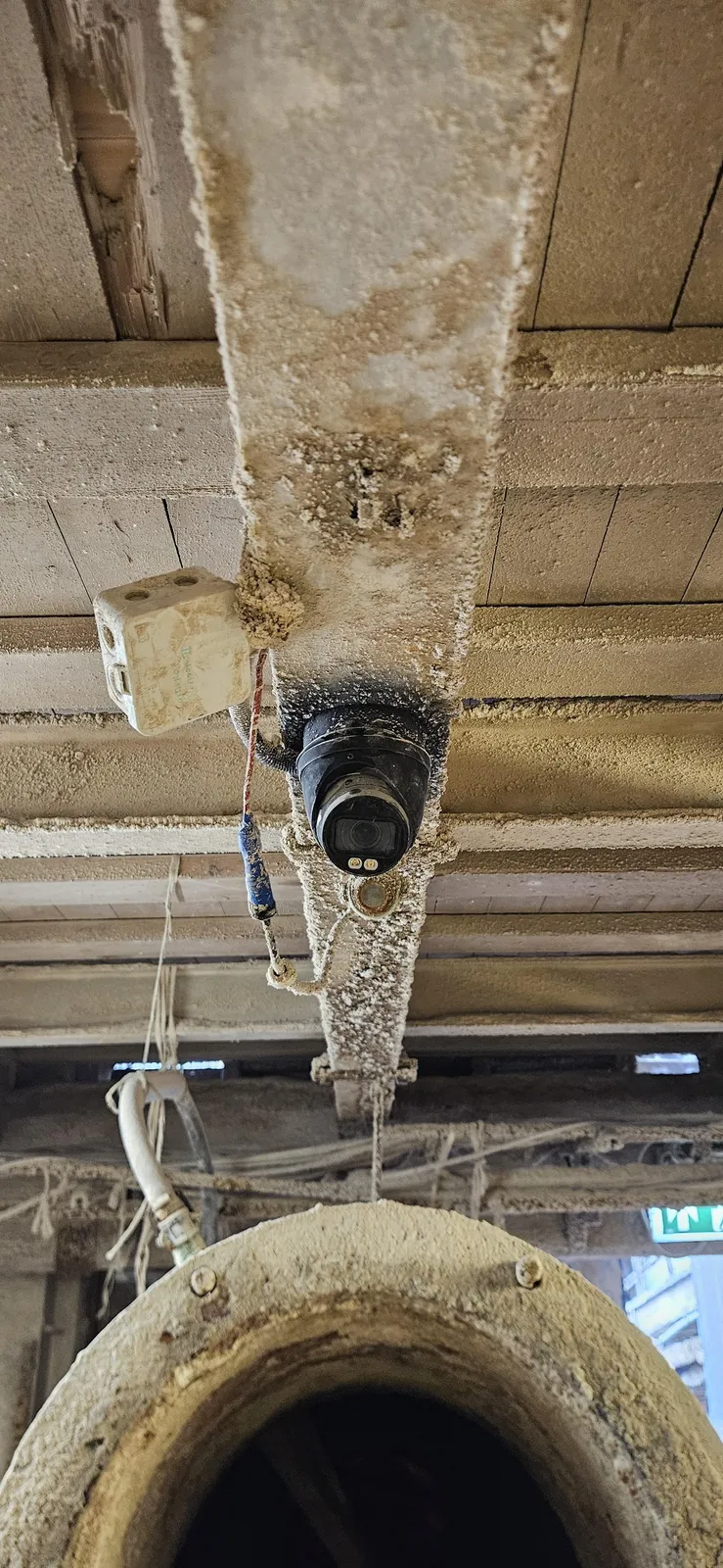 Zwart gecoate dome-camera onder een betonnen balk die dik bedekt is met fosfaatafzetting, boven een grote leiding in de fabriek