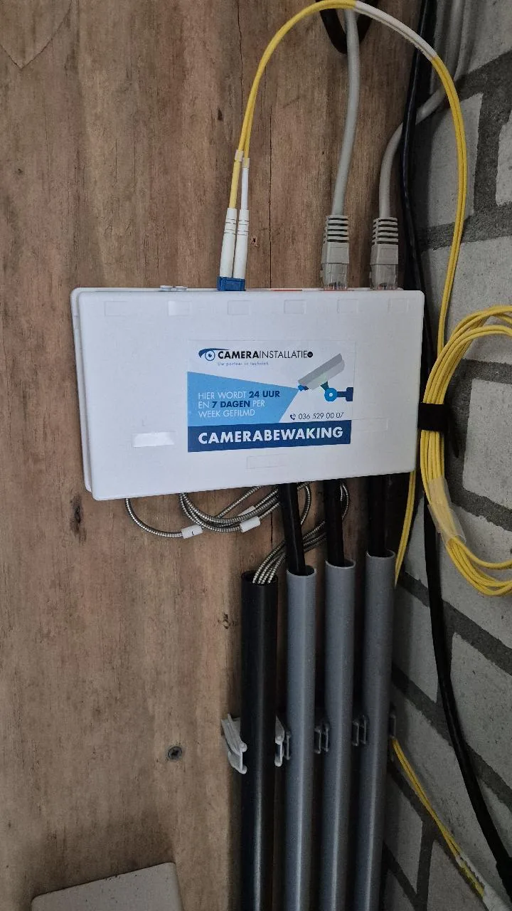 Gele glasvezelkabels en netwerkkabels bij een kast met de sticker van CameraInstallatie.nl in een technisch hokje