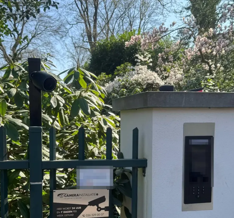 Intercom met scherm en cijfertoetsen ingebouwd in een witte pilaar naast de poort, met een camera op de hekpaal en een bord camerabewaking