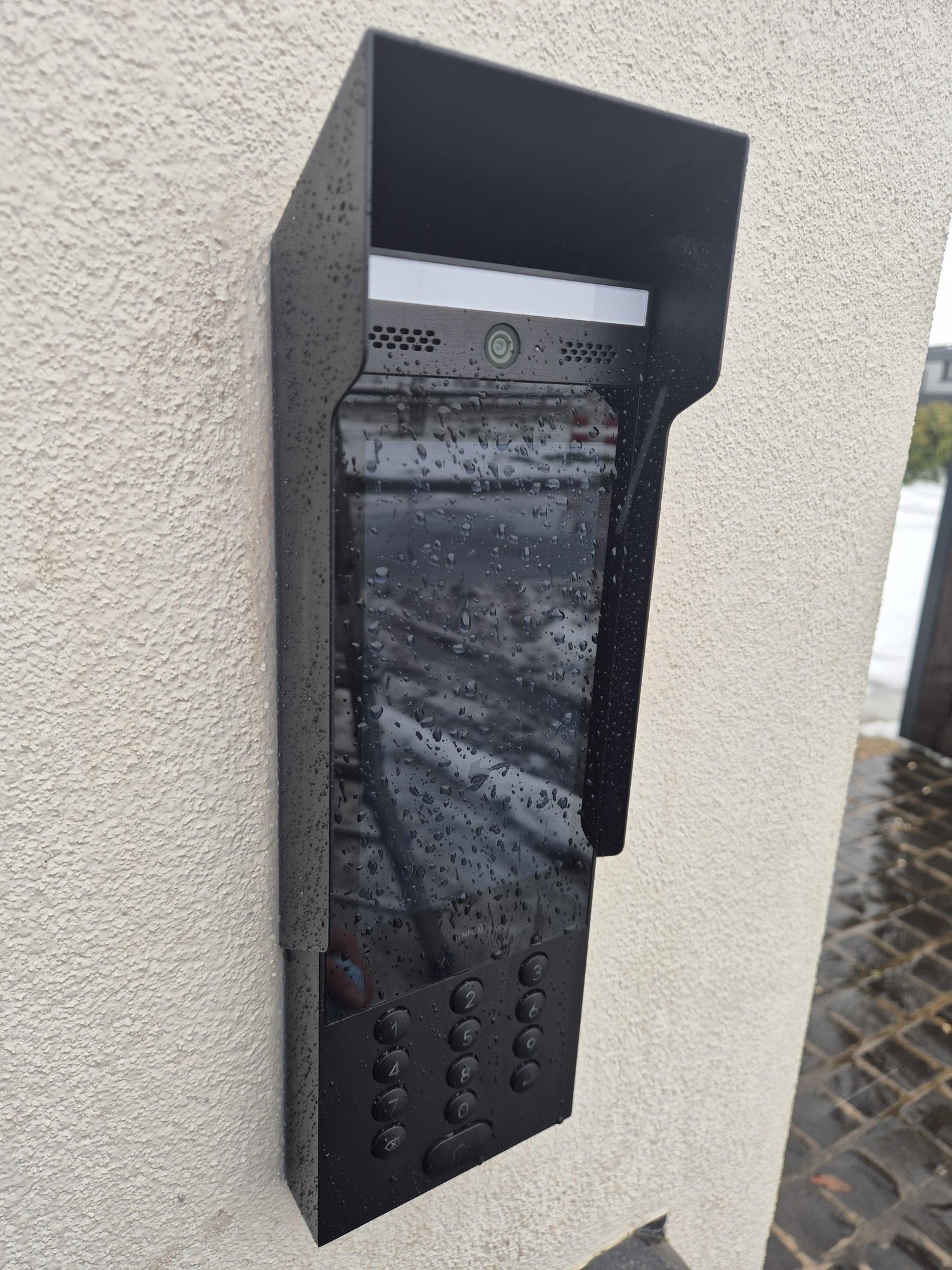 Zwarte intercom met camera, scherm en cijfertoetsen onder een kap, met regendruppels op het glas