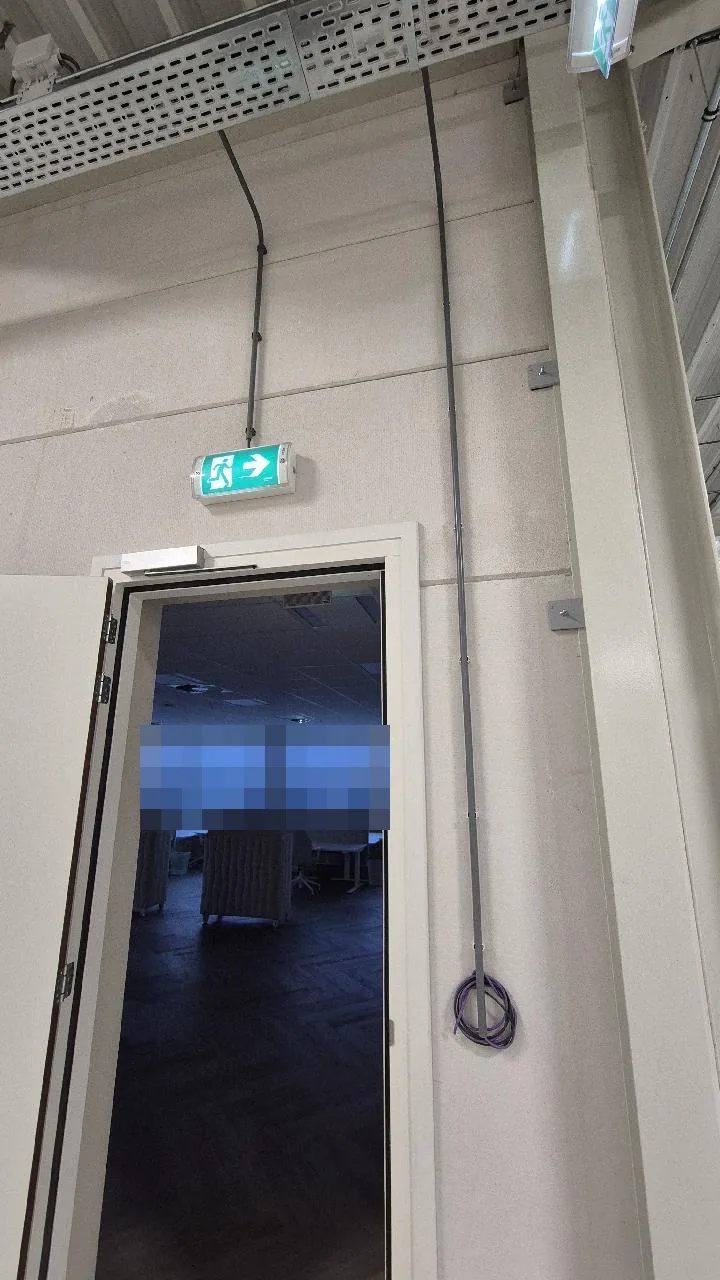 Kabel in grijze buis vanuit de kabelgoot langs de wand naar beneden naast een deur, met een opgerolde datakabel onderaan