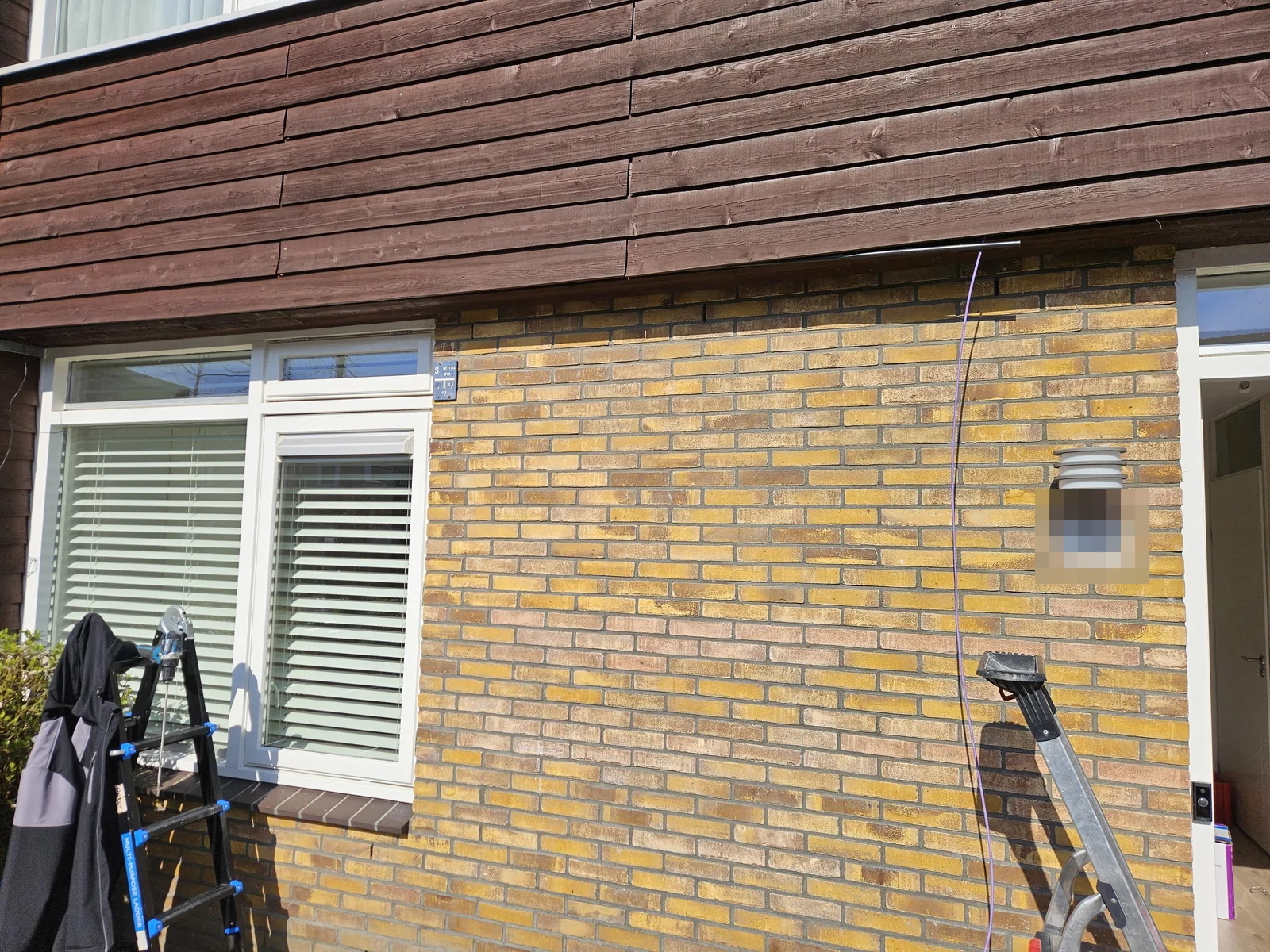 Gevel met ladders tijdens het werk, een kabel hangt langs de muur en een buis gaat onder de houten betimmering in