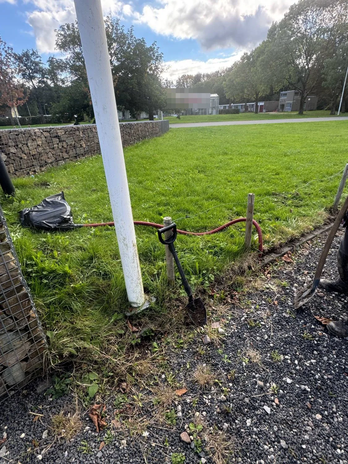 Open sleuf met een rode flexbuis in het gras, langs een vlaggenmast en een schanskorfmuur