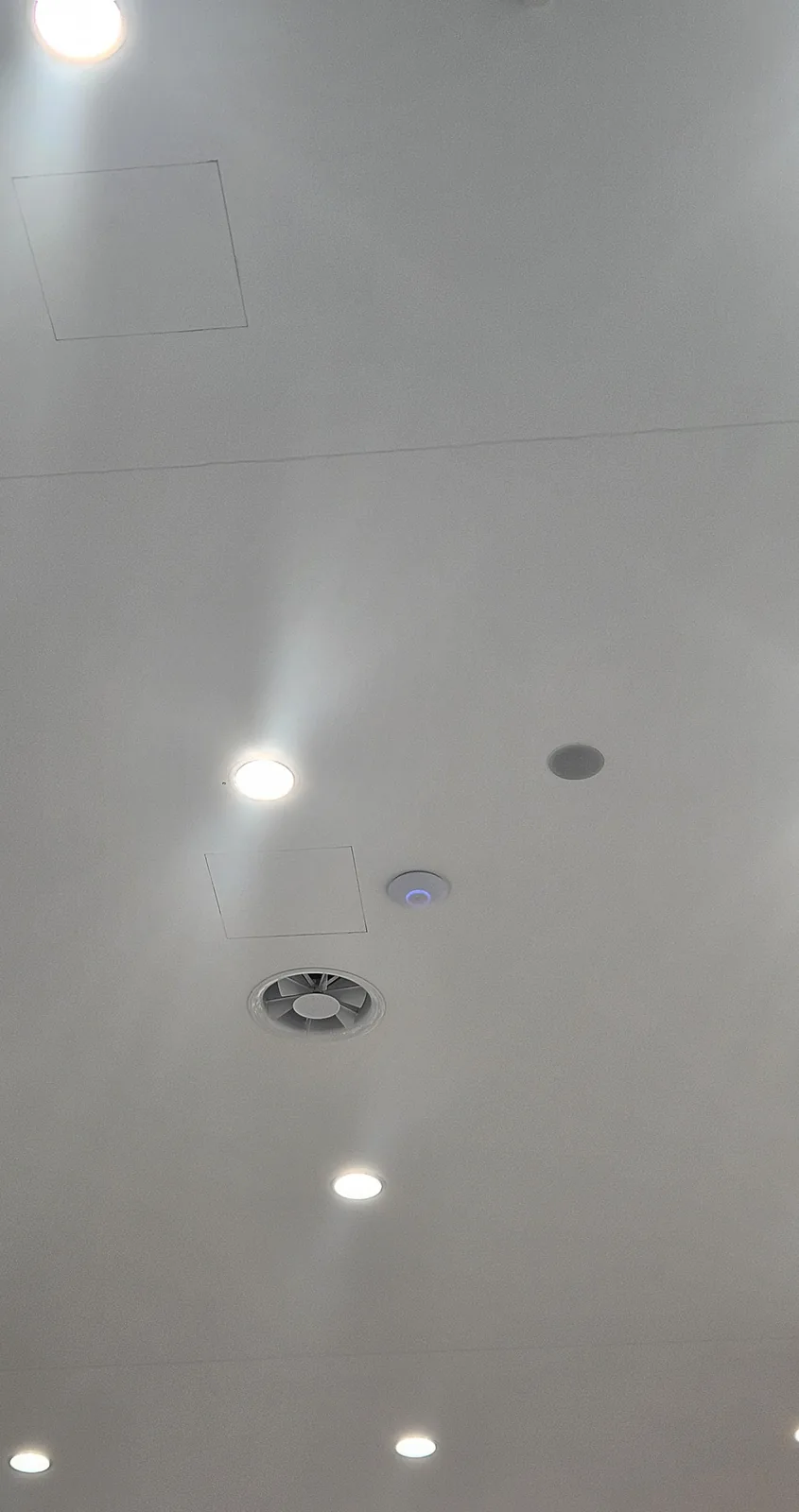 Lang wit kantoorplafond met inbouwspots en ventilatieroosters, met in het midden een access point met blauw lampje